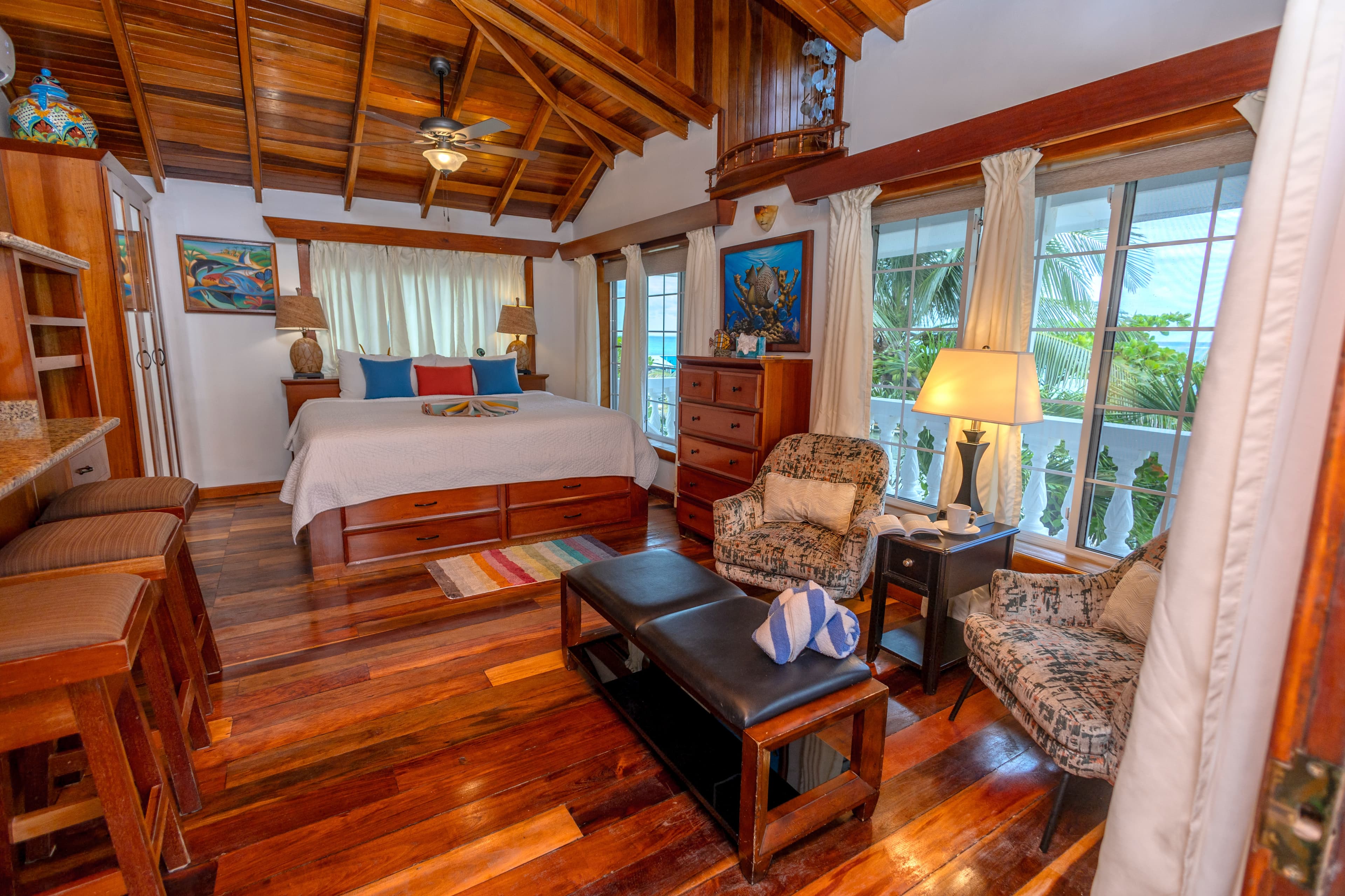 A bedroom with wood paneled walls and ceiling, a bed with colorful pillows, seating area, and balcony doors with sheer curtains.