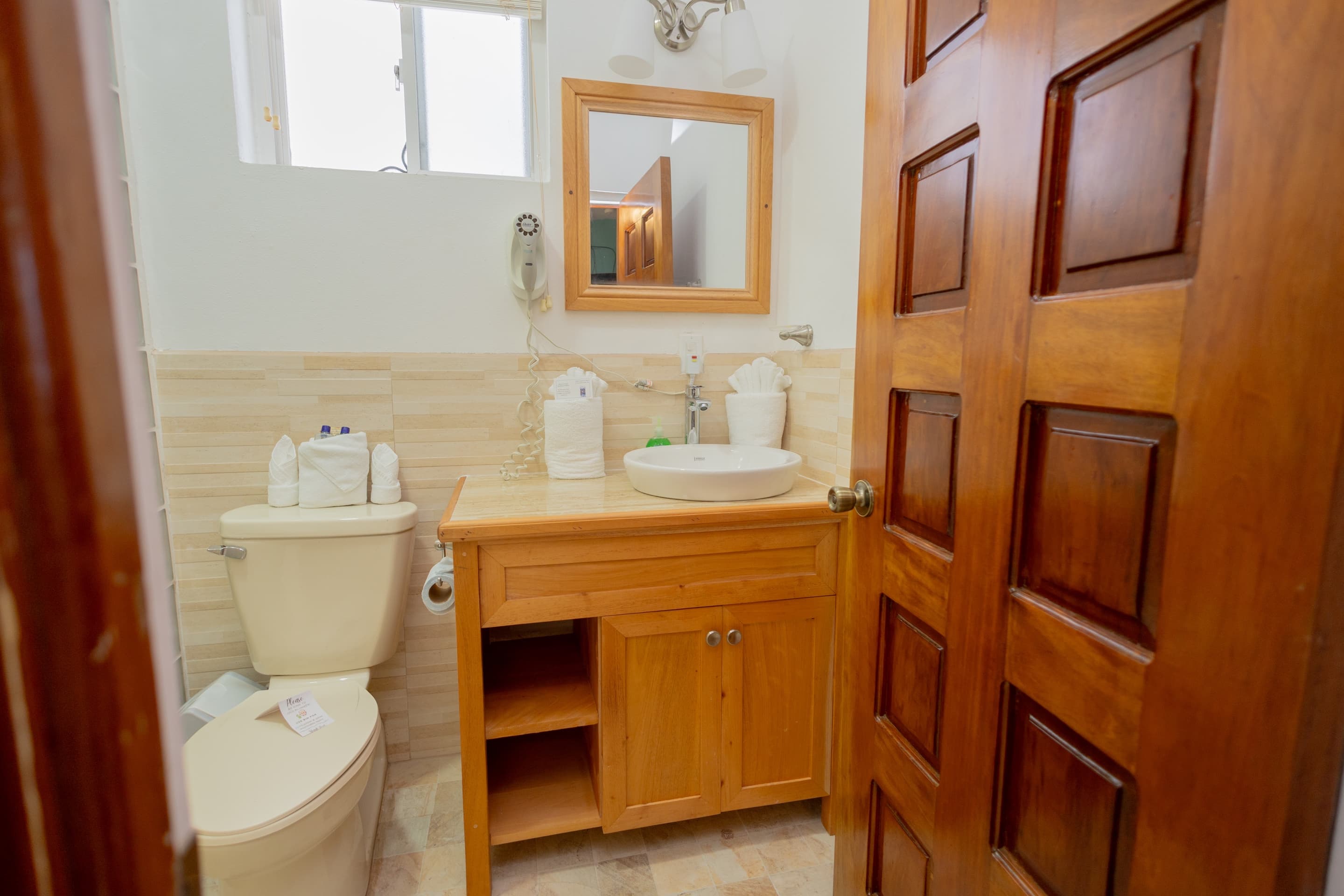 A compact bathroom with light tiled walls, a wooden-framed mirror above a sink with a wooden cabinet, and a white toilet, viewed through a partially open wooden door.