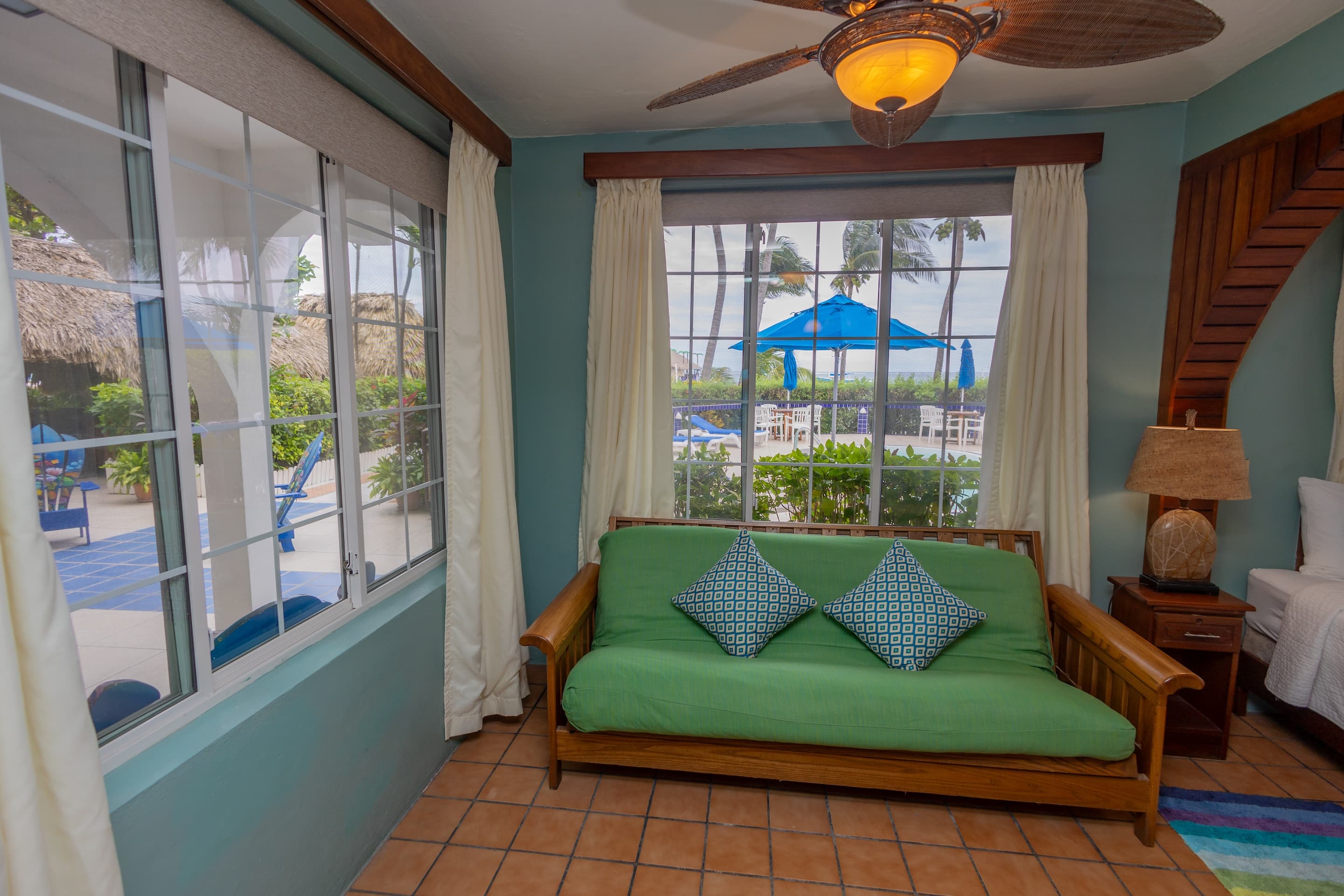 A room with teal walls, tiled floors, a green futon with blue pillows, a ceiling fan, and windows with a view of a pool area.