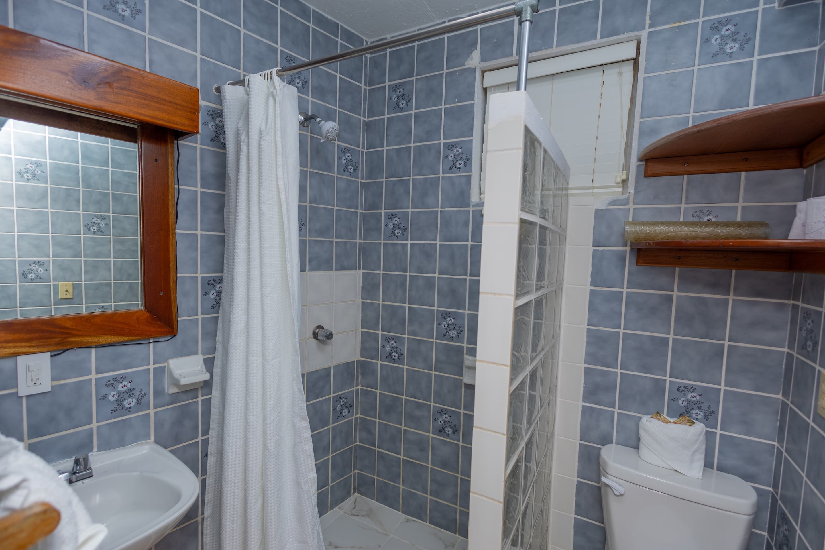 A compact bathroom with blue and gray patterned tiles, a wooden-framed mirror above a white sink, a shower with a white curtain, and a toilet.