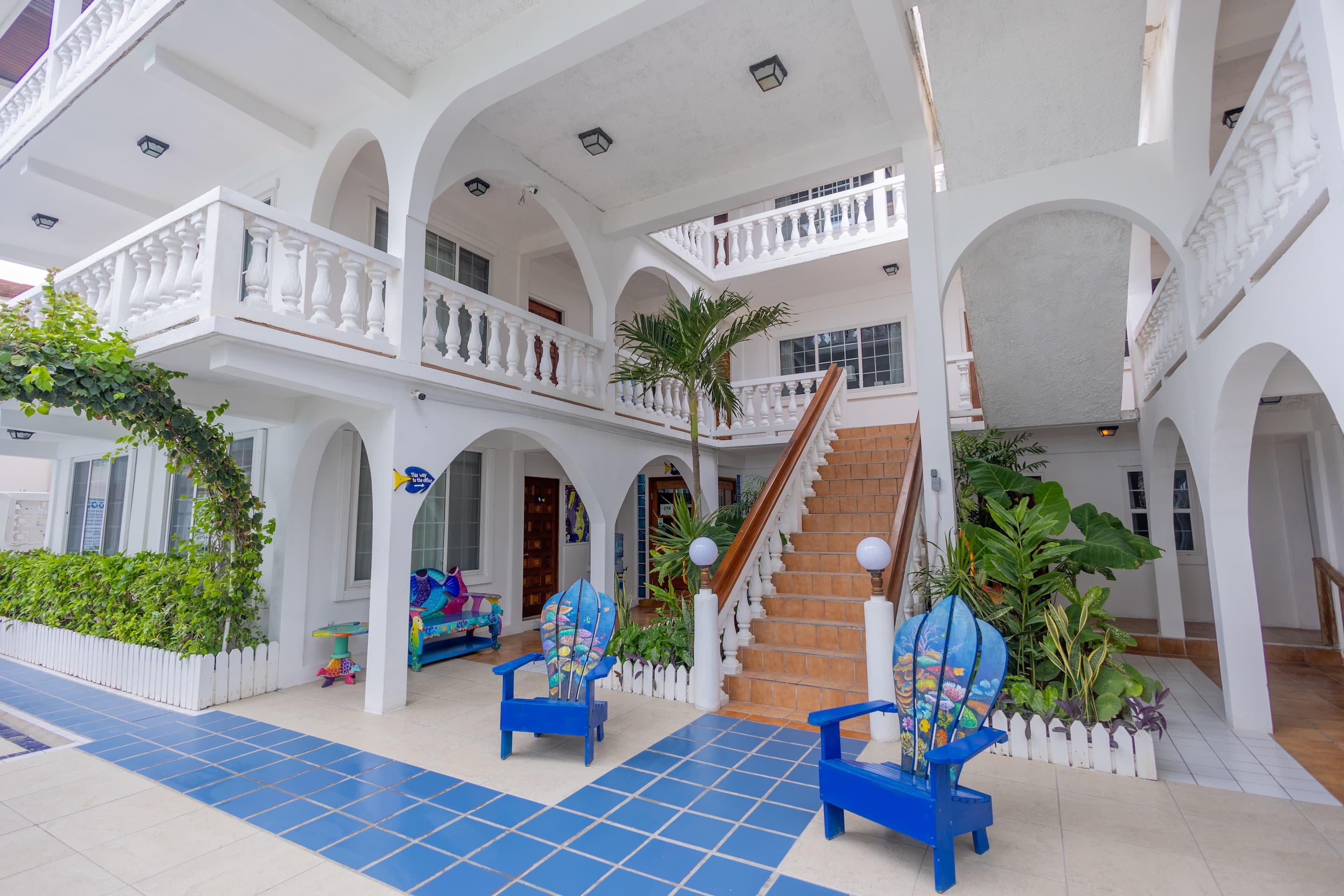 A white courtyard with blue tiled walkways, lush greenery, and blue Adirondack chairs.