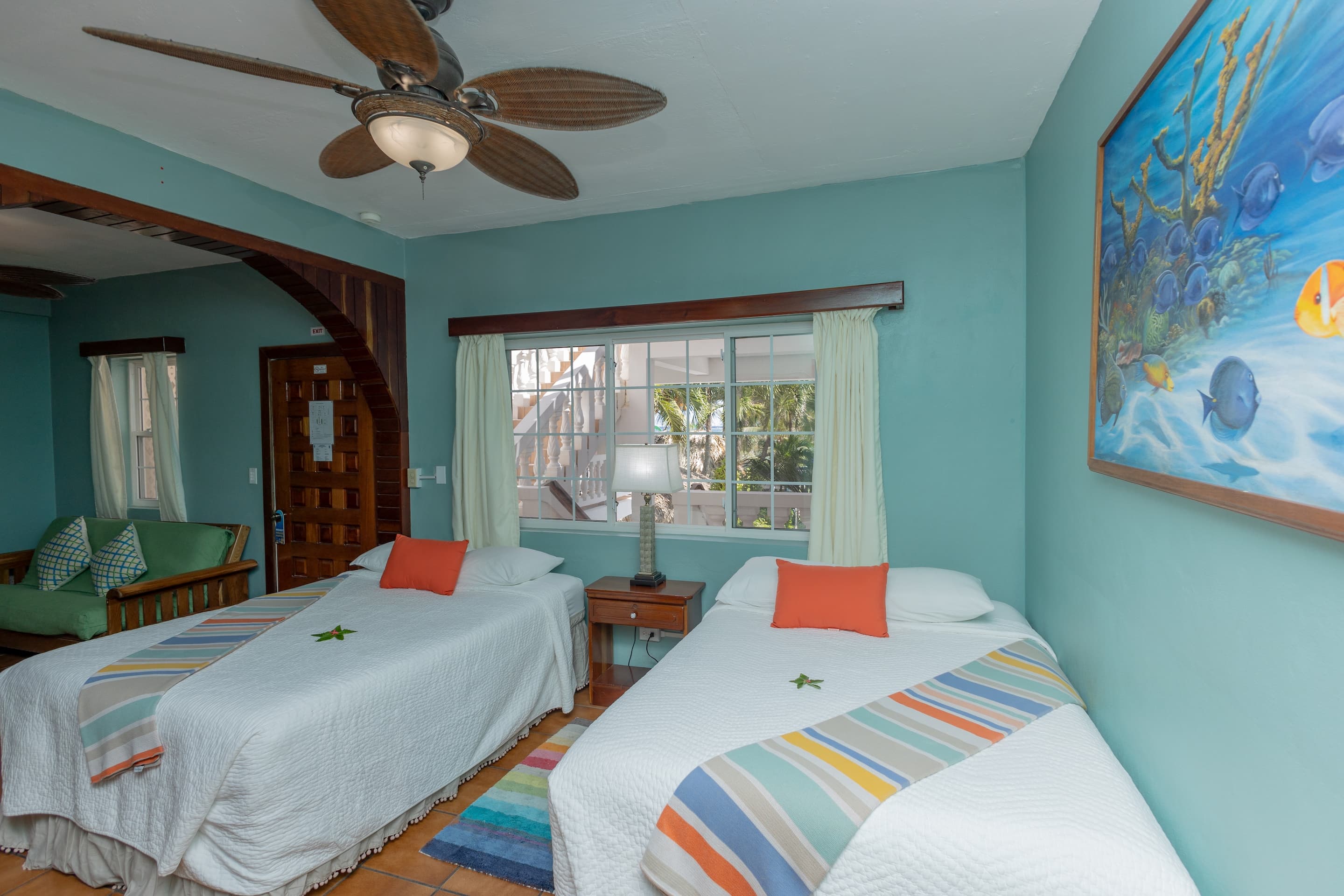 A bedroom with teal walls, tiled floors, two full beds with colorful accents, and a ceiling fan.
