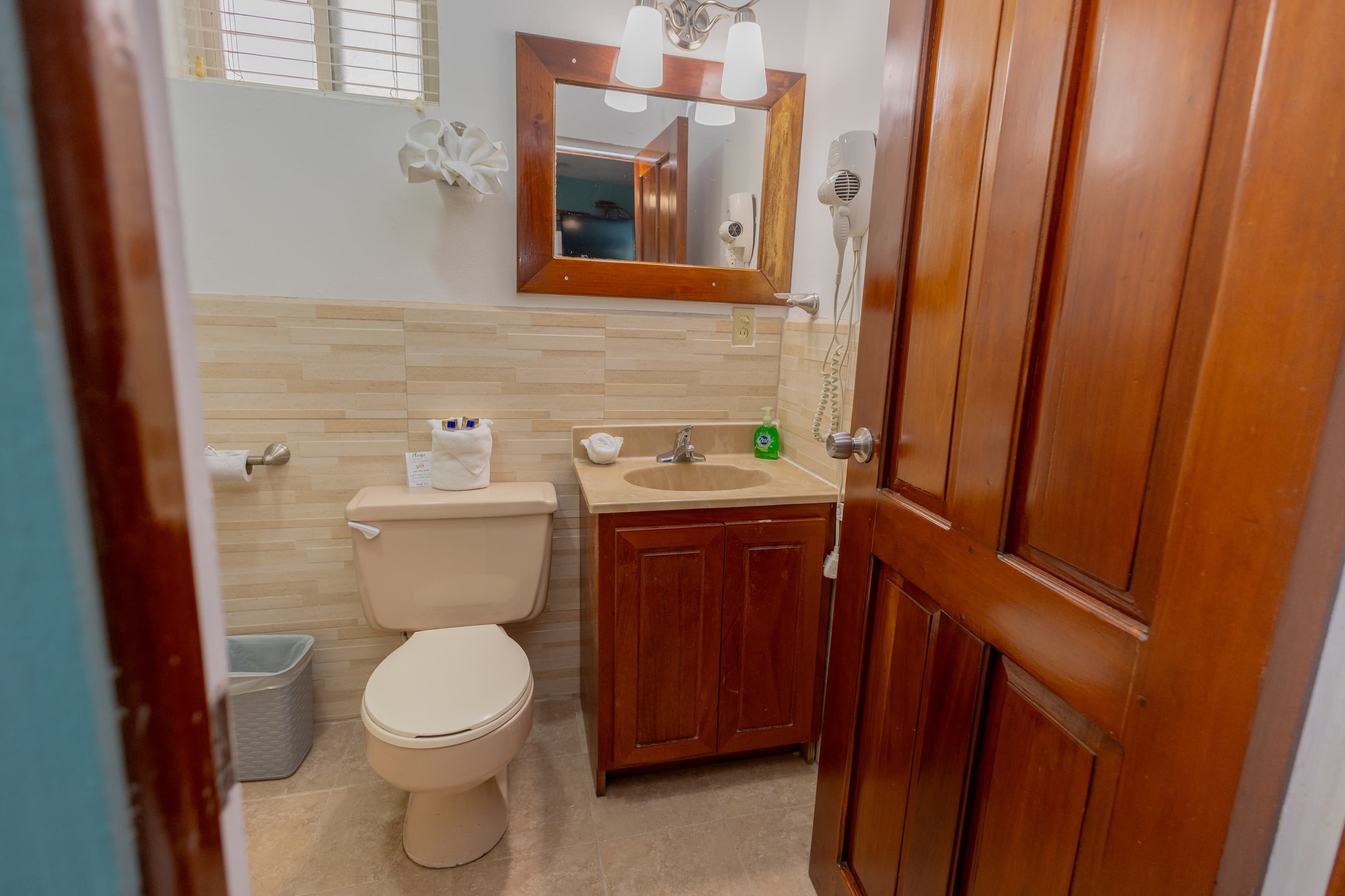 A compact bathroom with light tiled walls, a wooden-framed mirror above a sink with a wooden cabinet, and a white toilet, viewed through a partially open wooden door.