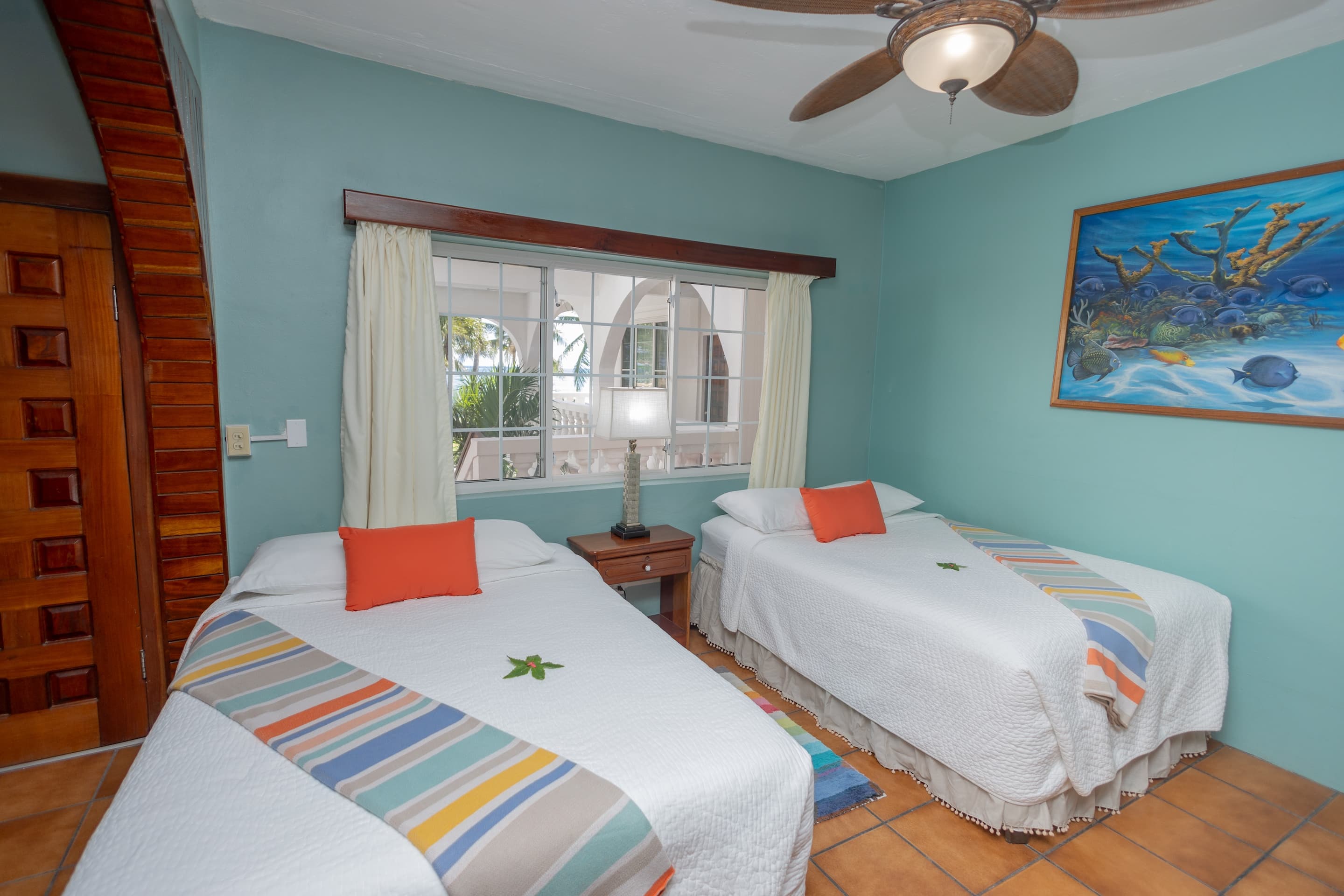 A bedroom with teal walls, tiled floors, two full beds with colorful accents, and a ceiling fan.