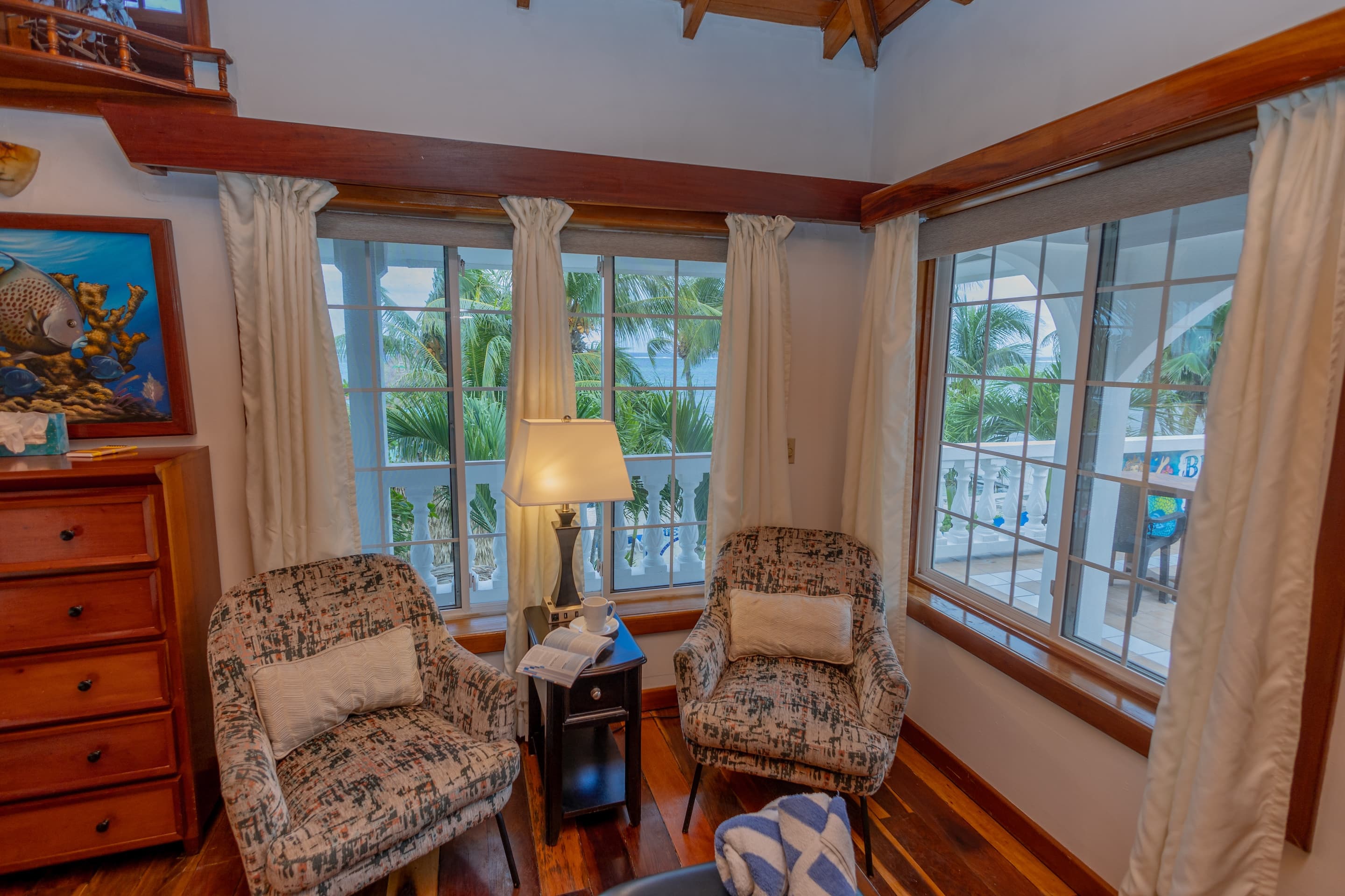 A sitting area with two patterned armchairs, a small table with a lamp, and windows with sheer curtains letting in natural light.