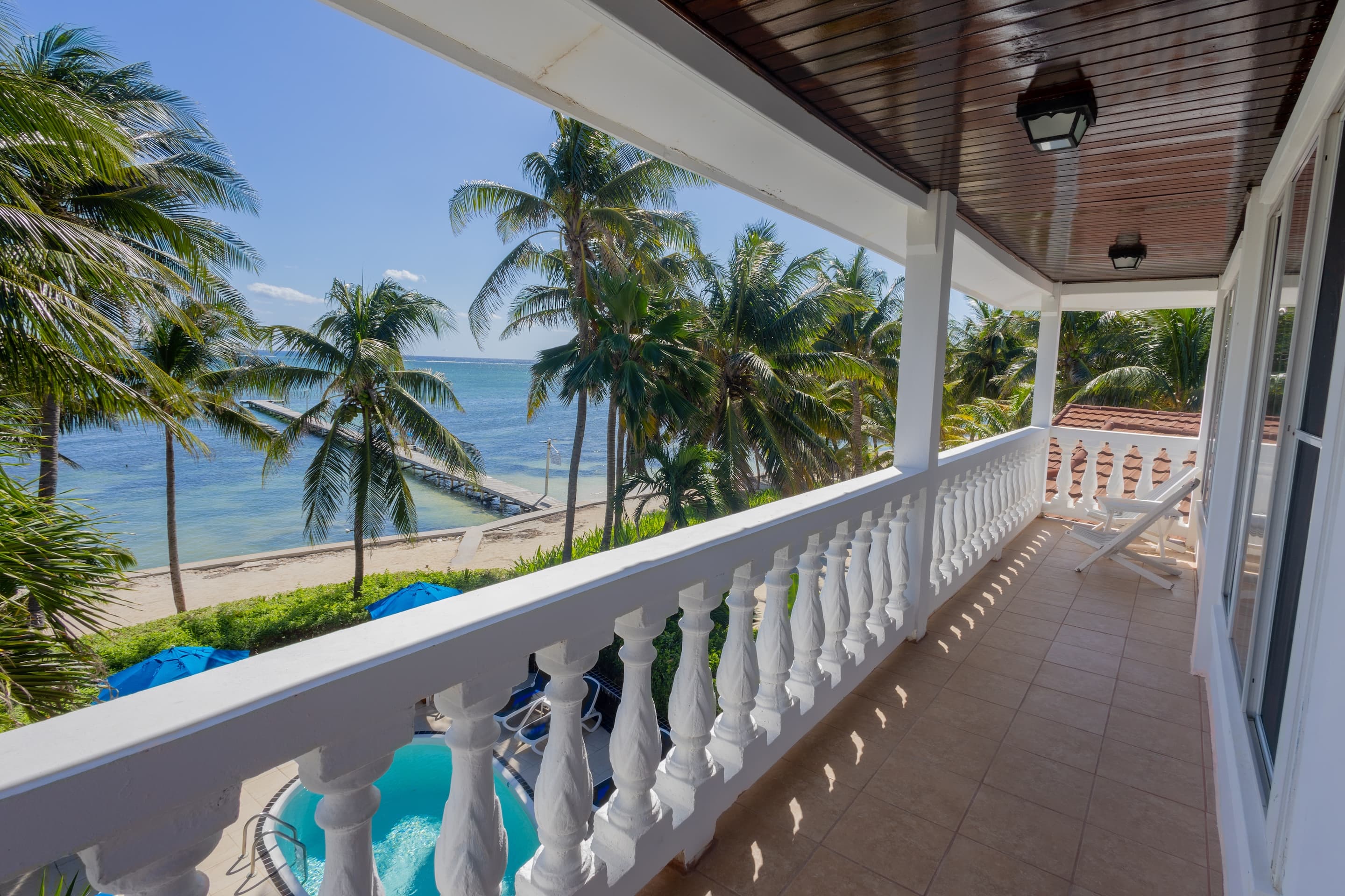 A long, covered balcony with white balustrades and a tiled floor, offering a scenic view of a tropical beach.