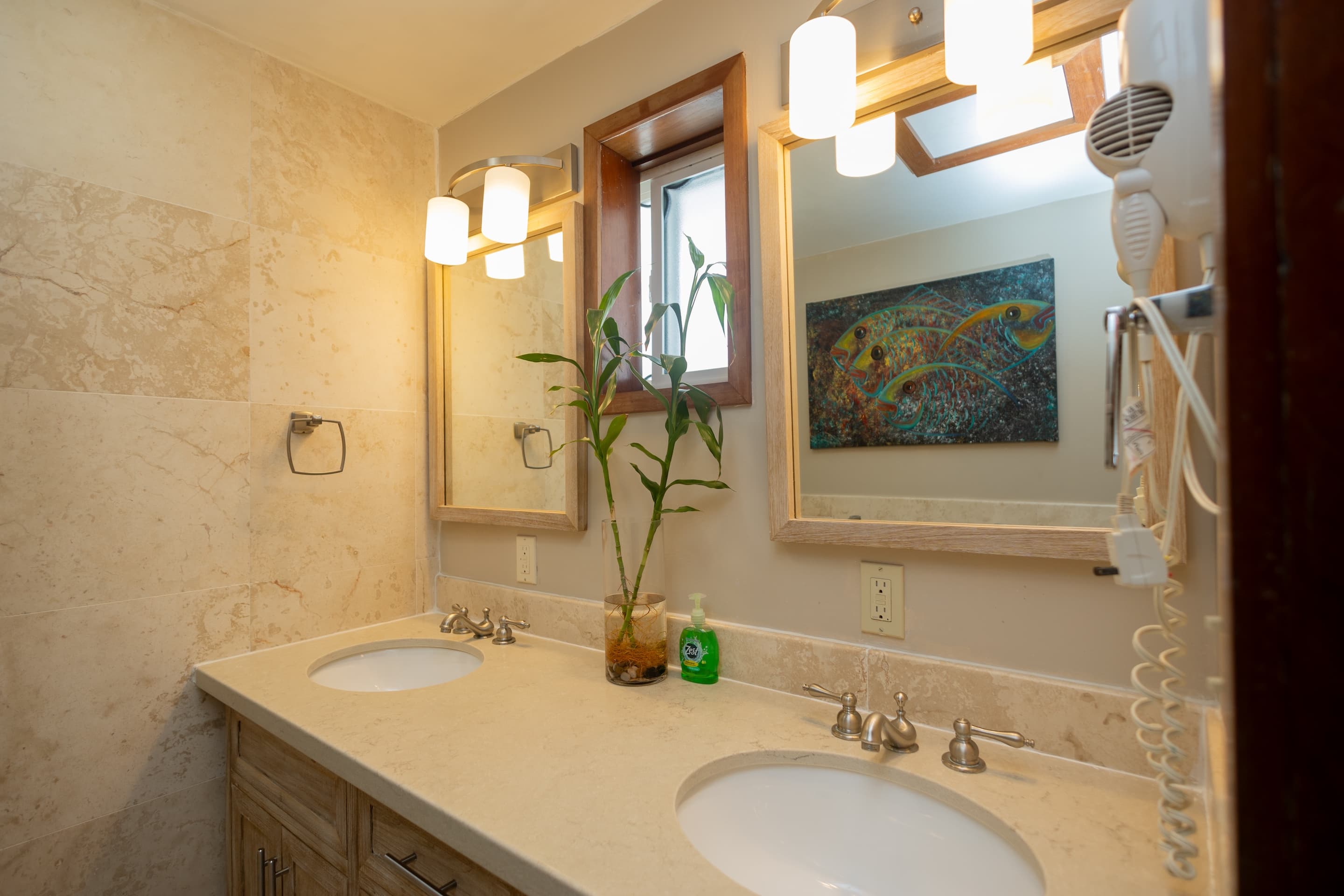 A bathroom featuring a double vanity with light countertops and white sinks, wooden-framed mirrors with overhead lighting, and light marble-patterned walls.