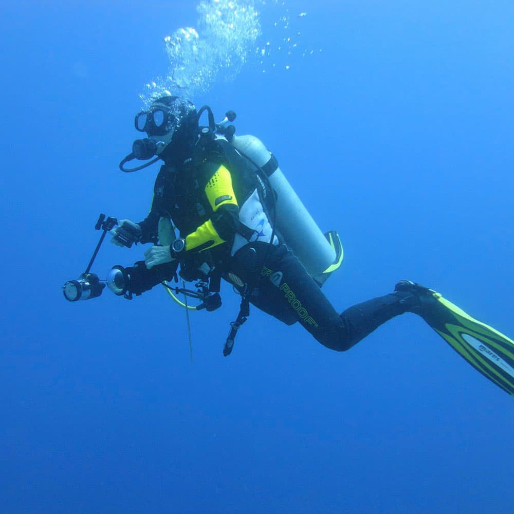 A scuba diver in a black wetsuit and yellow sleeves maneuvers underwater with a camera setup.