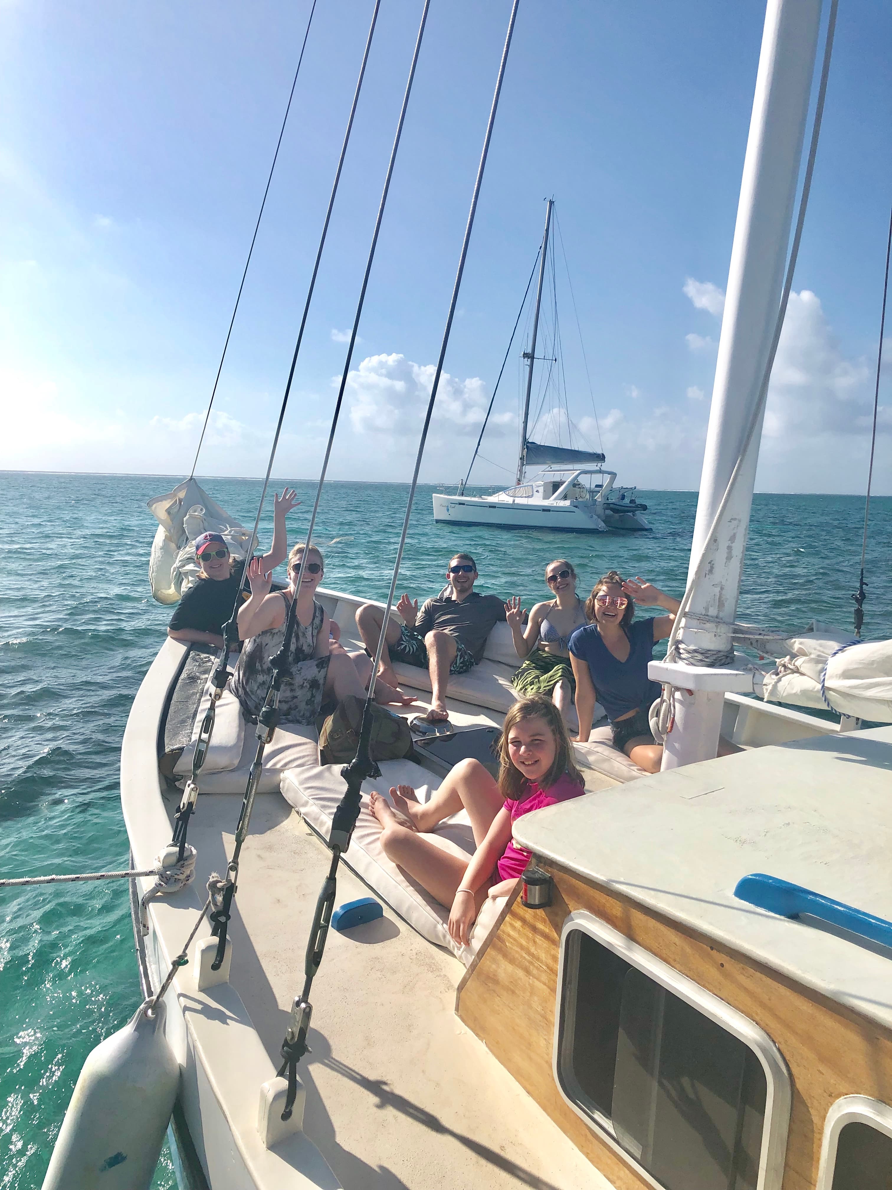 A group of seven people happily relax on a sailboat in clear blue waters under a sunny sky.