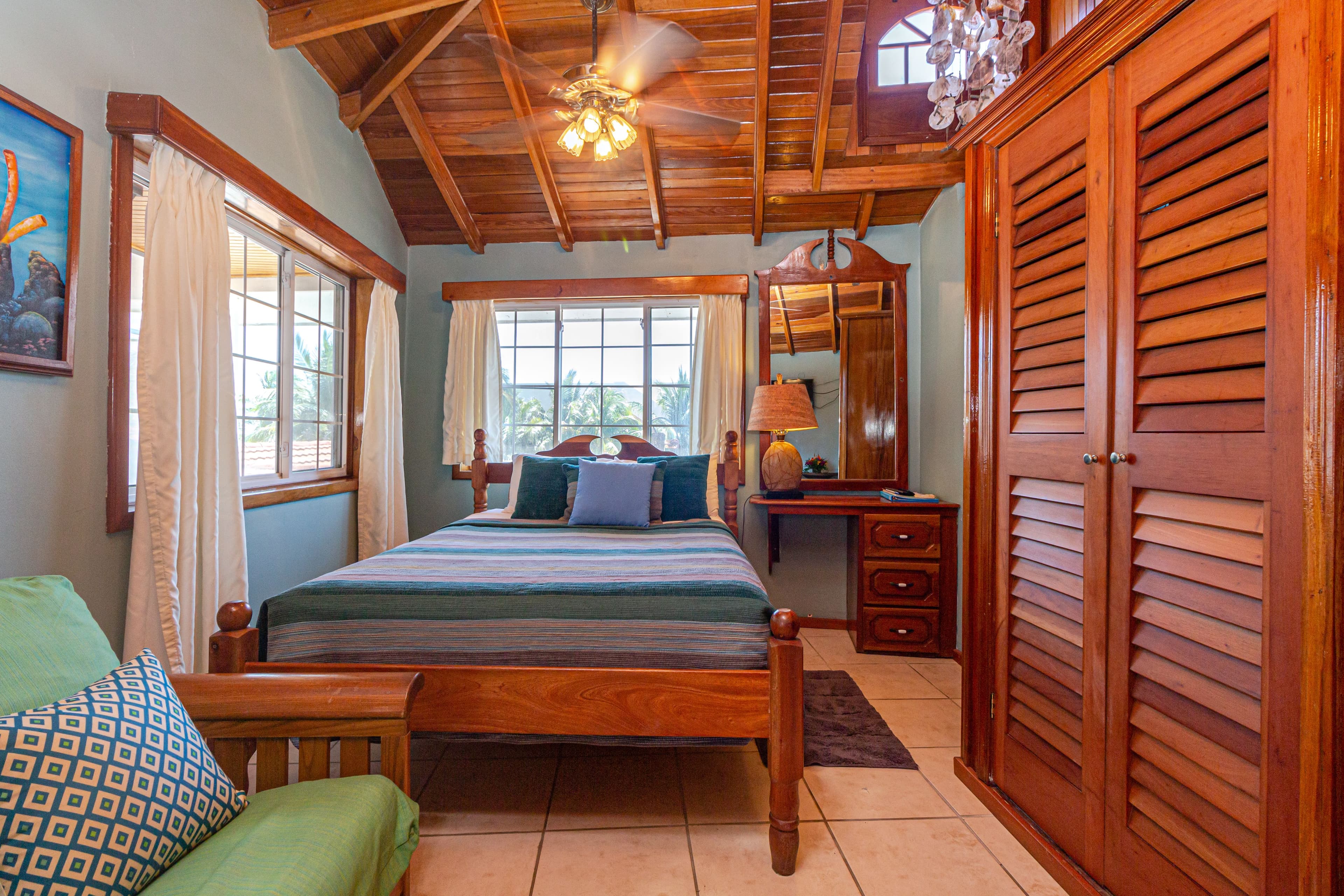 A room with a high wooden ceiling, a queen bed with blue bedding, a green futon, and a wooden wardrobe.