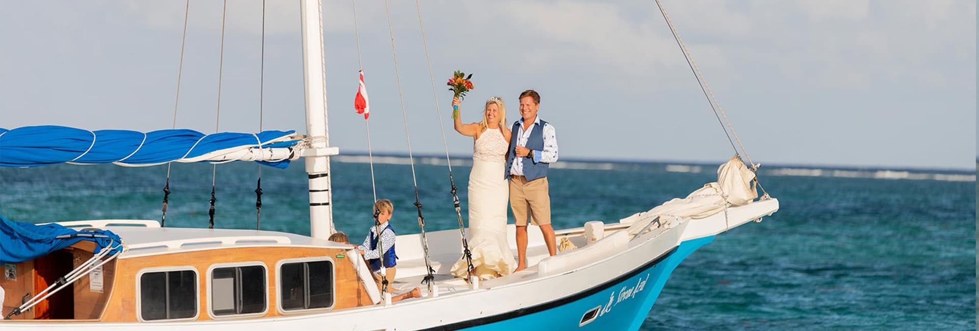 A bride and groom celebrate their wedding on the bow of a boat with a child sitting nearby.
