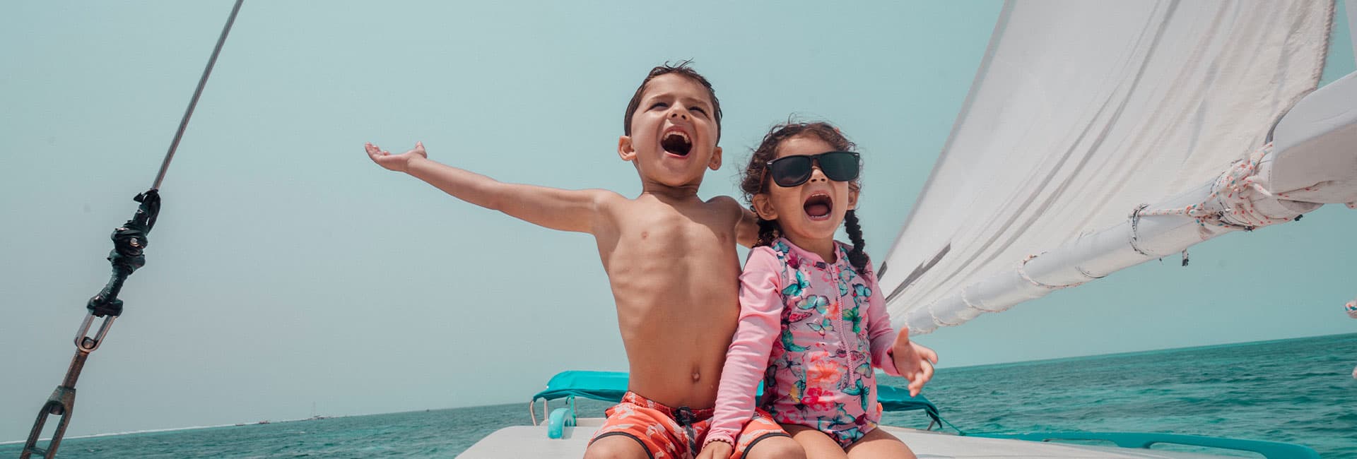 Two excited children, one shirtless boy and one girl in a swimsuit, joyfully shout and pose on a boat, surrounded by water and a clear sky.