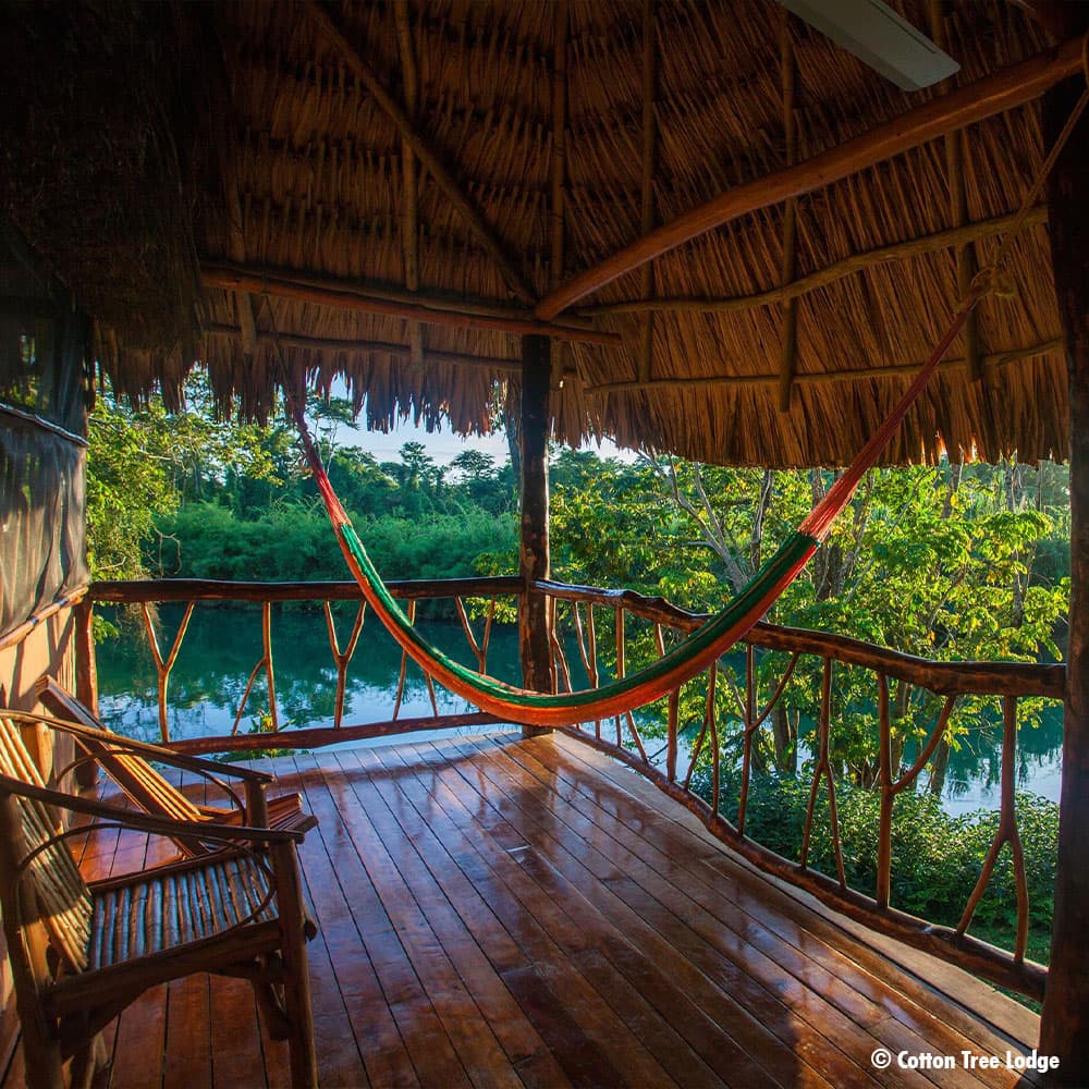 A wooden balcony with a hammock overlooks a lush green landscape and a tranquil river.