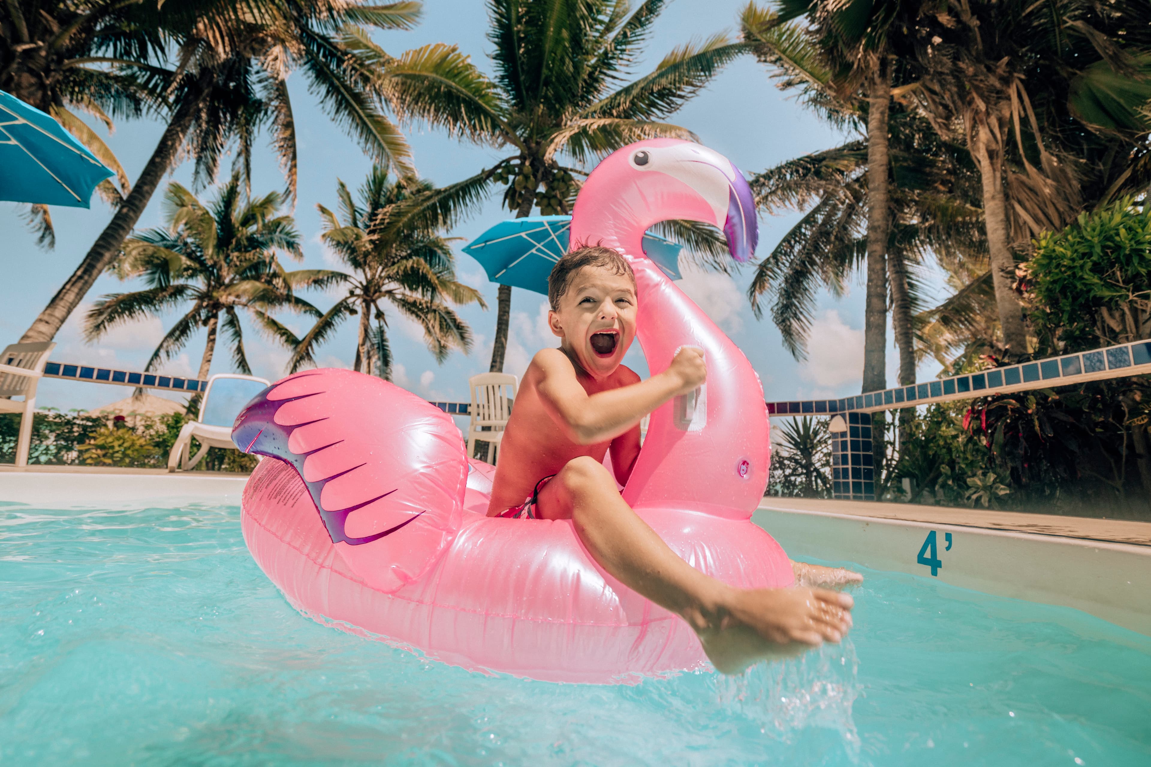 A joyful child splashes in a pool on a pink flamingo float, surrounded by palm trees and bright sunshine.