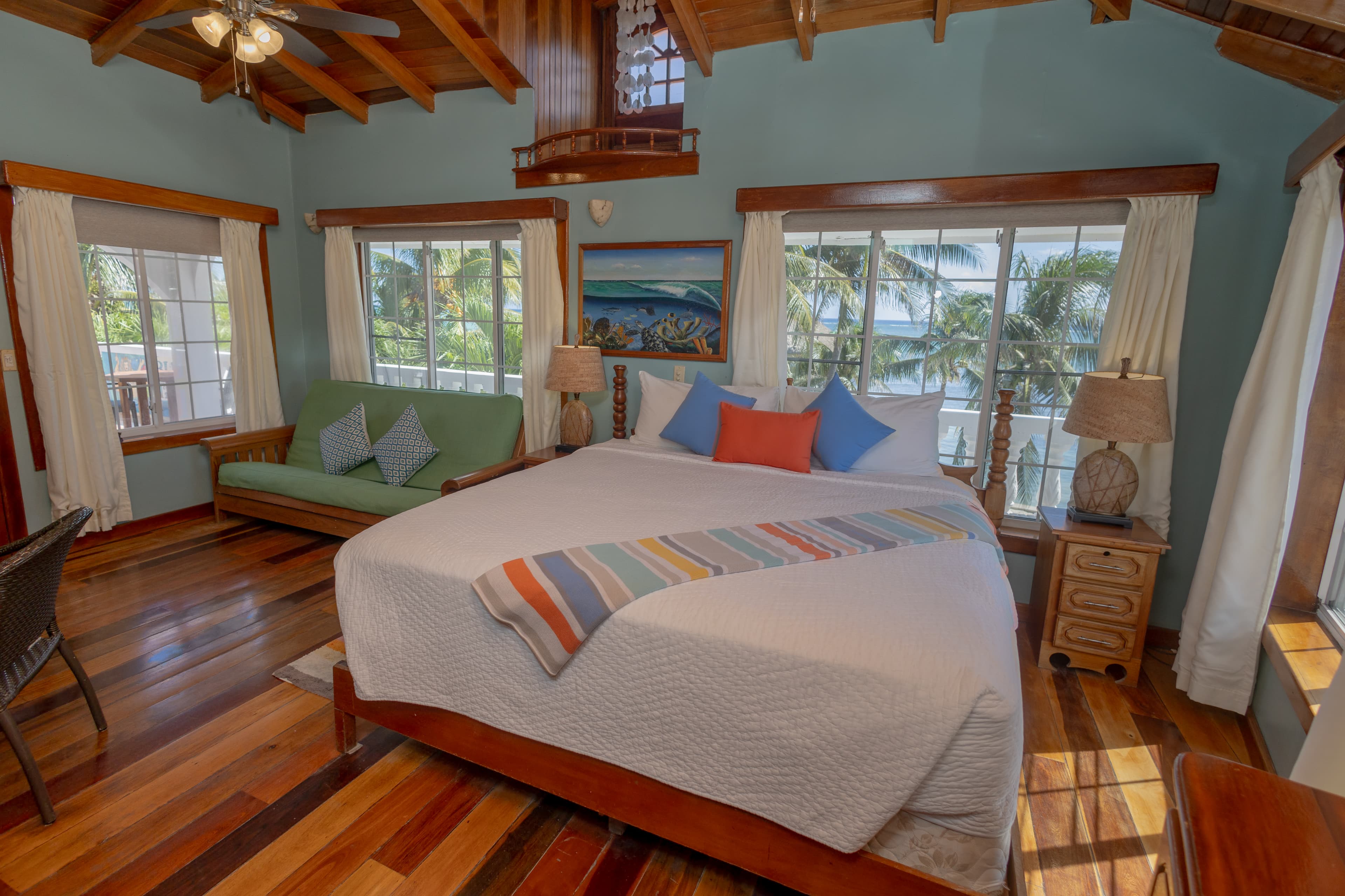 A cozy bedroom with a large bed, green couch, and windows overlooking palm trees.