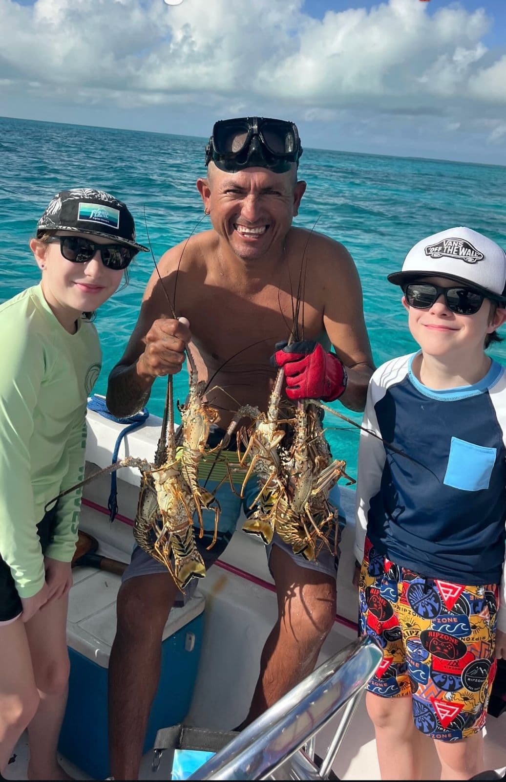 A smiling man holds several lobsters while posing with two children on a boat in clear blue waters.