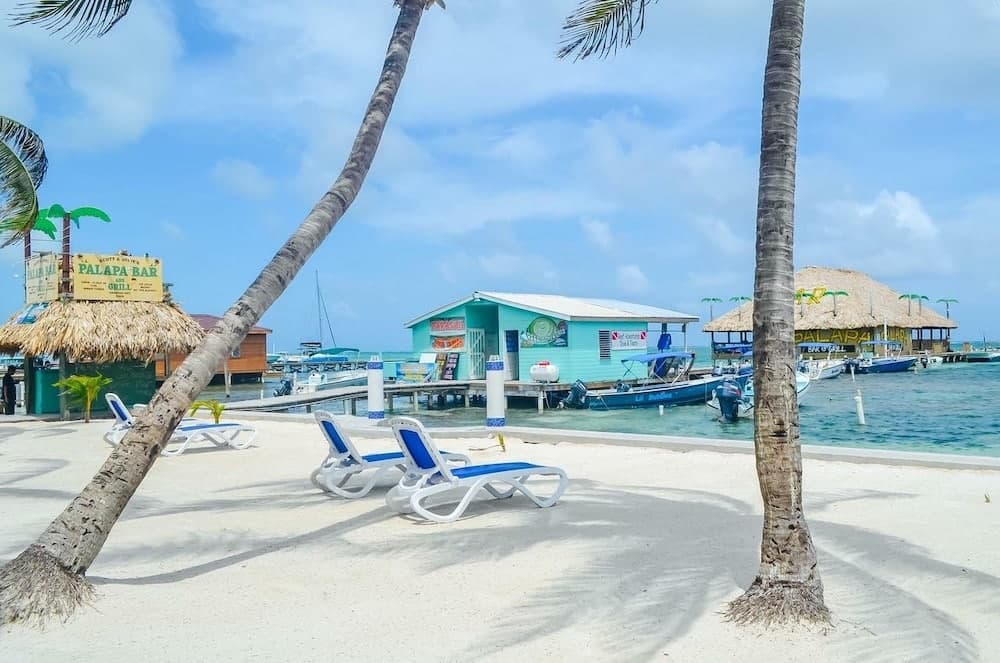 A scenic beach view with loungers, palm trees, and colorful seaside buildings.