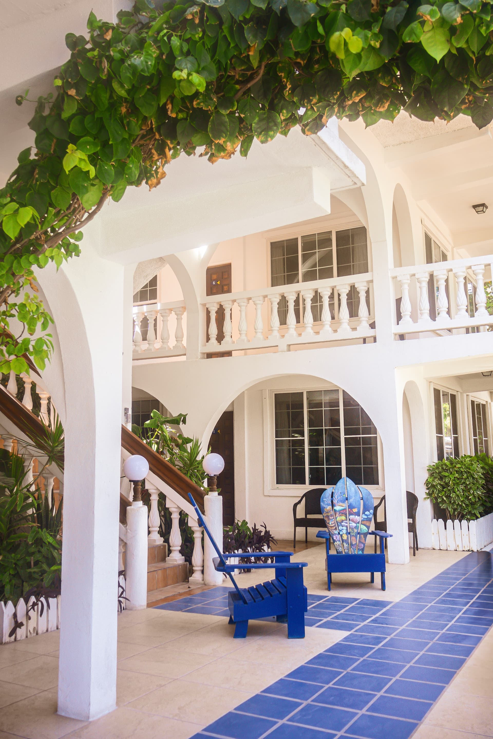 A white courtyard with blue tiled walkways, lush greenery, and blue Adirondack chairs under a vine-covered archway.
