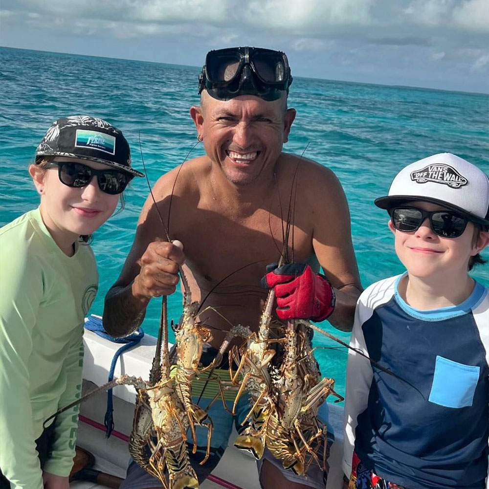 A man and two children, all smiling, hold freshly caught lobsters against a turquoise sea backdrop.