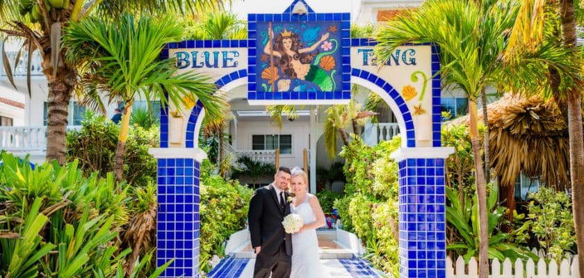 A couple poses happily under a colorful tiled archway labeled "BLUE TING" surrounded by lush greenery.