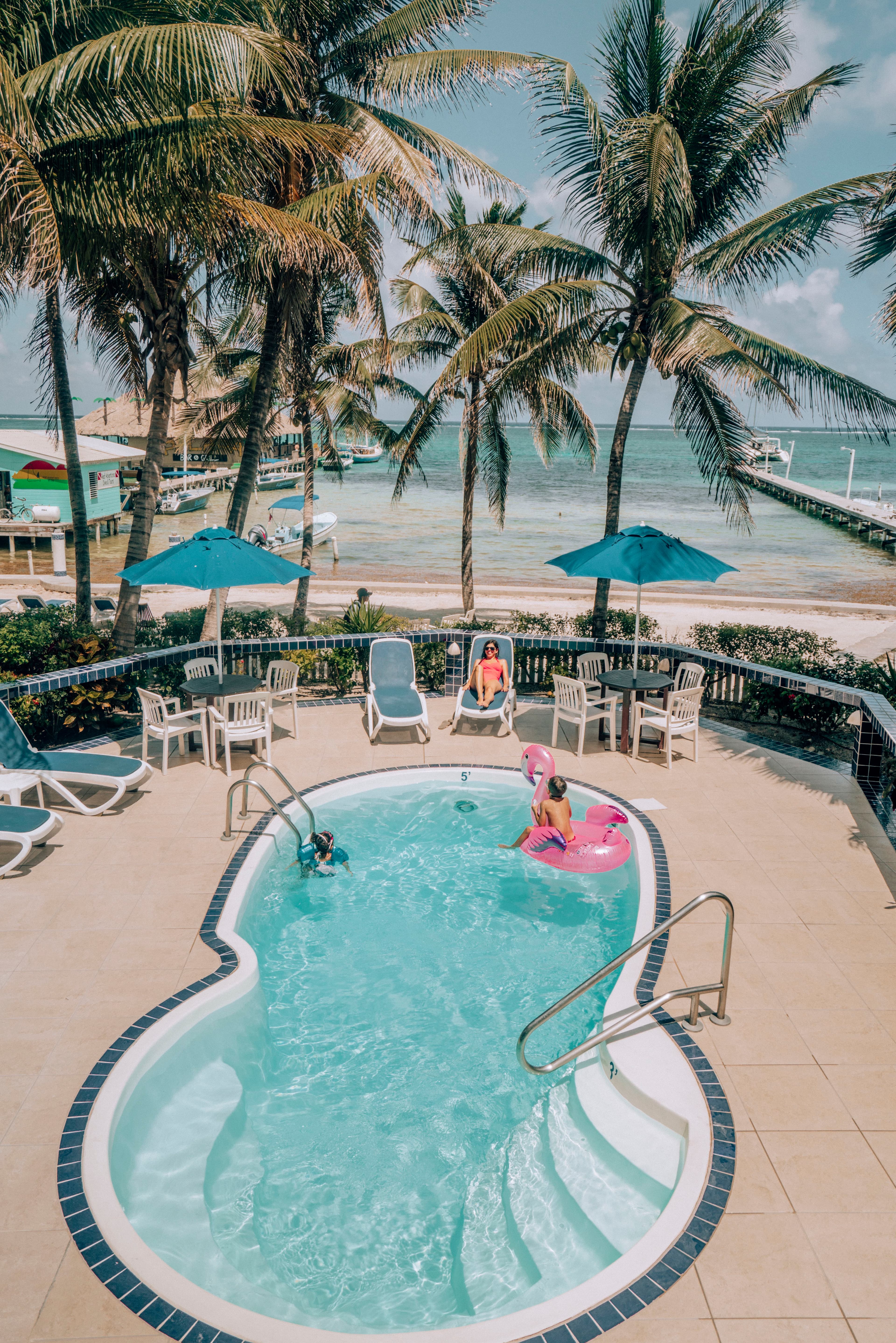 A sparkling pool surrounded by palm trees, with two children playing and a woman lounging nearby.