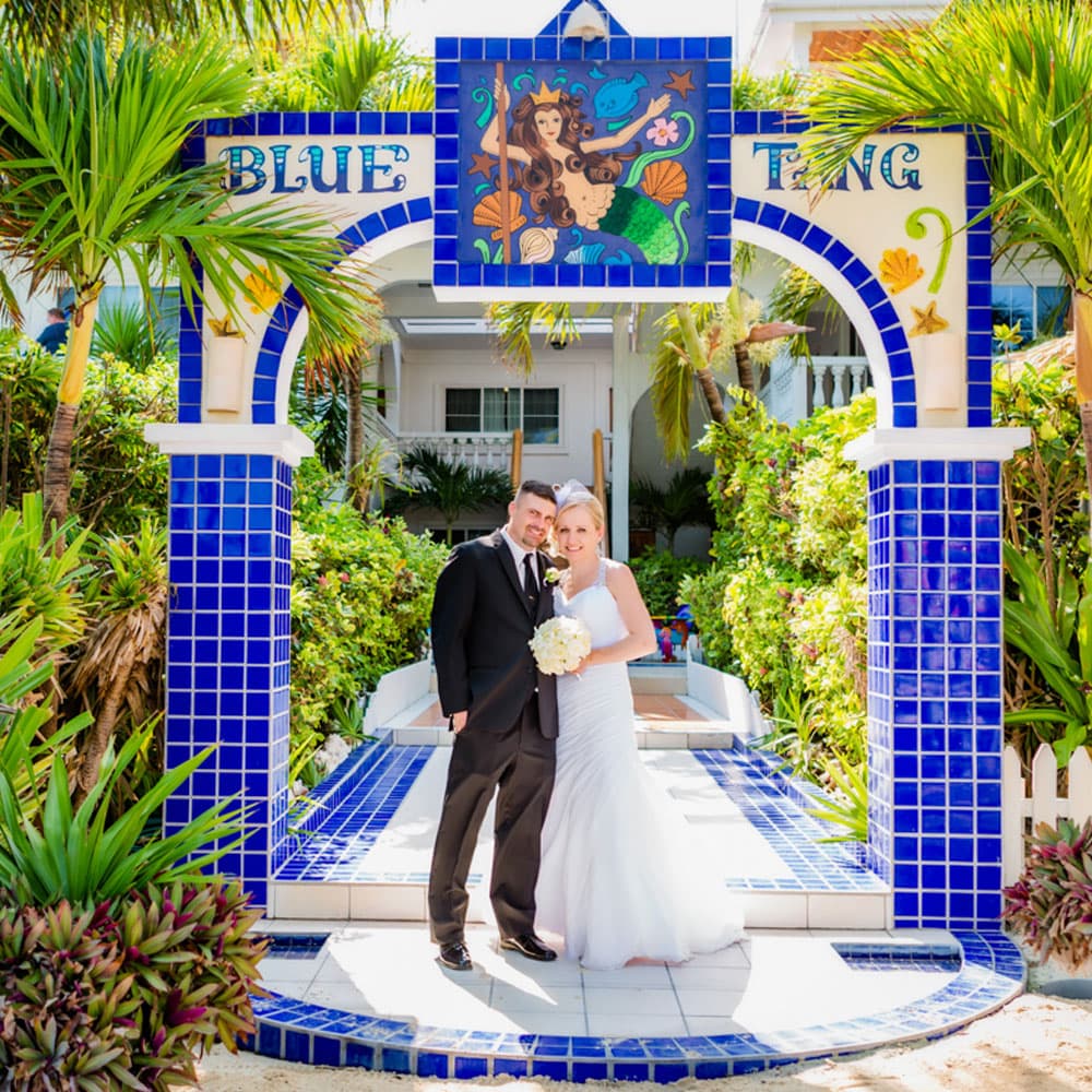 A smiling couple in wedding attire stands under a blue-tiled archway adorned with a mermaid mural.