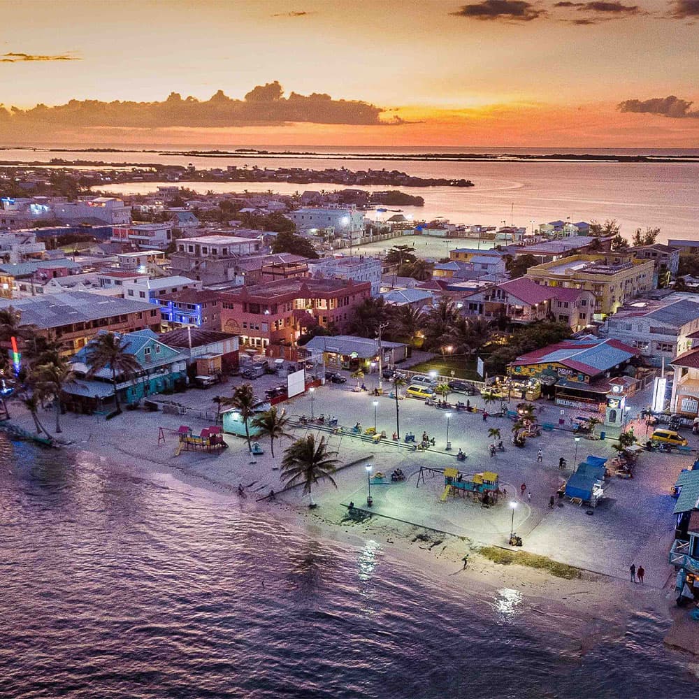 Aerial view of a coastal town at sunset with colorful buildings, palm trees, and a sandy beach.