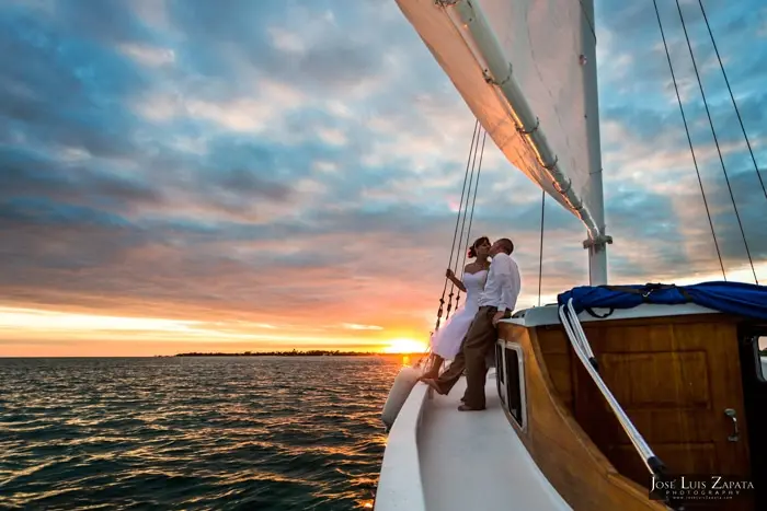A couple embraces on the bow of a sailboat during a sunset.