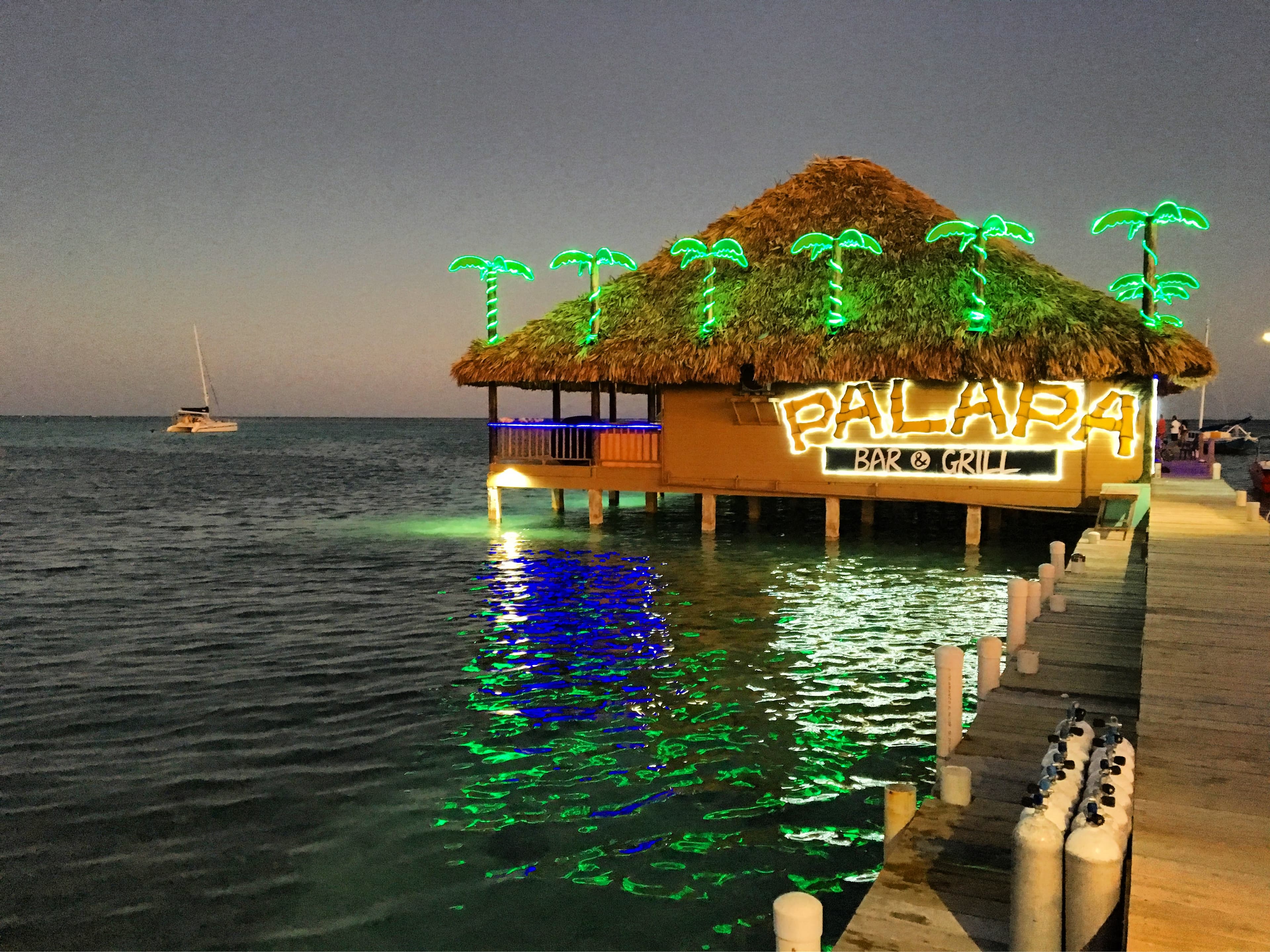 A lit tiki bar with palm trees stands over water at dusk, with a boat in the background.