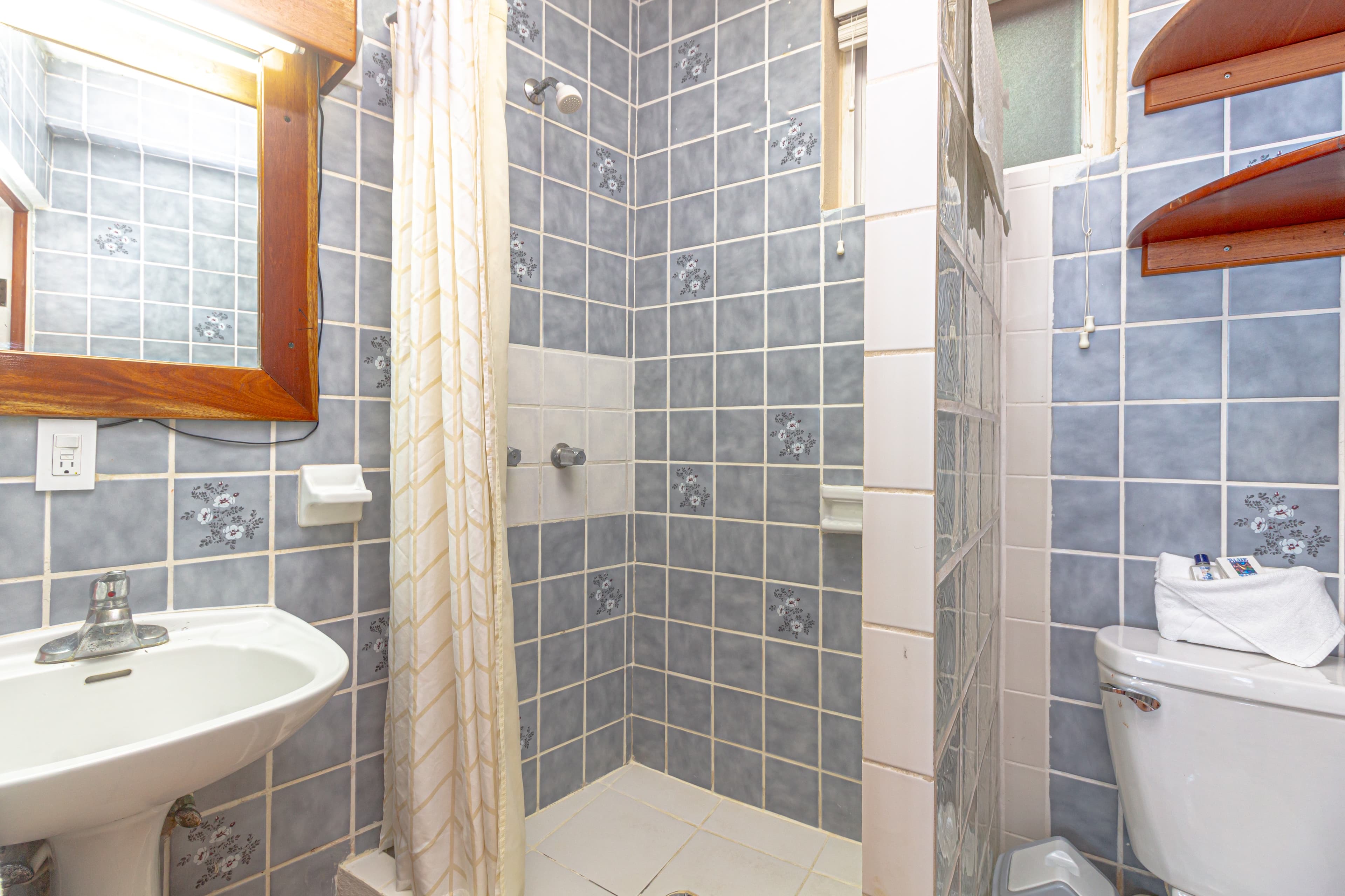 A compact bathroom with blue and gray patterned tiles, a wooden-framed mirror above a white sink, a shower with a white curtain, and a toilet.