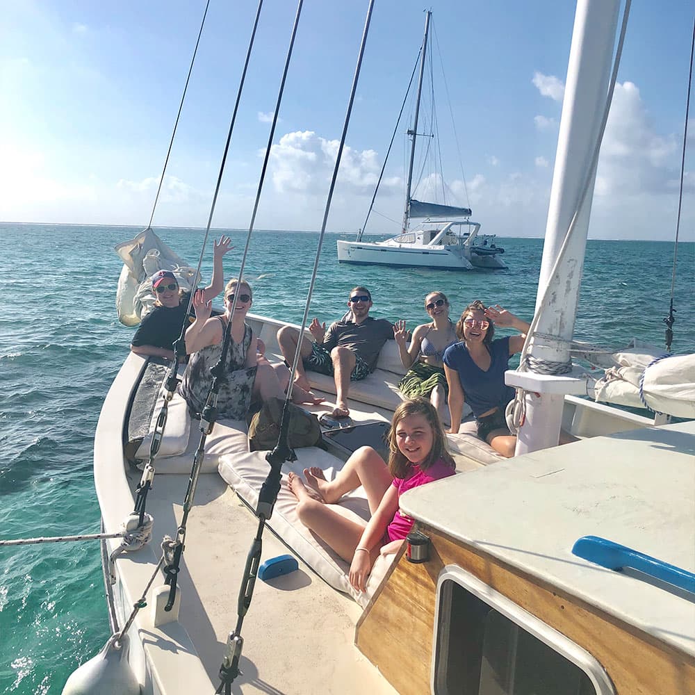 A group of six people are sitting and waving on the deck of a sailboat in clear blue waters.