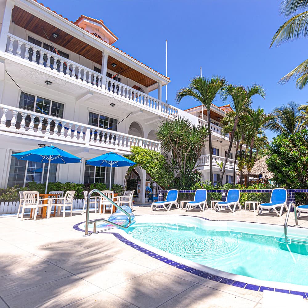 A sunny pool area with lounge chairs and blue umbrellas next to a white building surrounded by palm trees.
