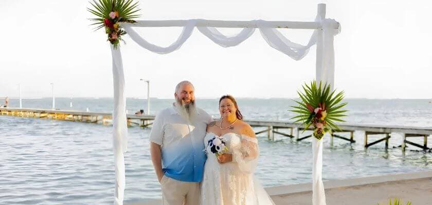 A smiling couple stands under a floral arch by the water, celebrating their wedding.