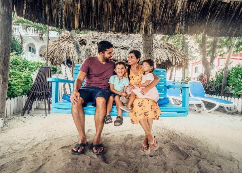 A smiling family of four sits on a blue swing under a thatched roof, enjoying a beach setting. A smiling family of four sits on a blue swing under a thatched roof, enjoying a beach setting.