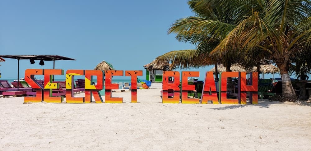 Colorful "Secret Beach" sign on a sandy beach with palm trees and lounge chairs in the background.