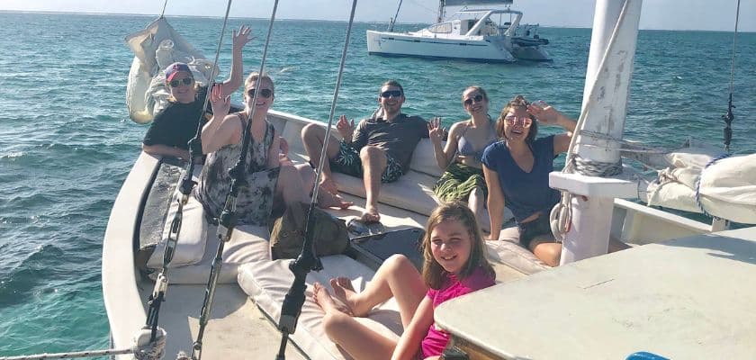 A group of six people on a sailboat waves happily at the camera while cruising on the water.