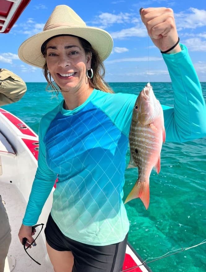A smiling woman in a sun hat holds a freshly caught fish while standing on a boat in clear blue water.
