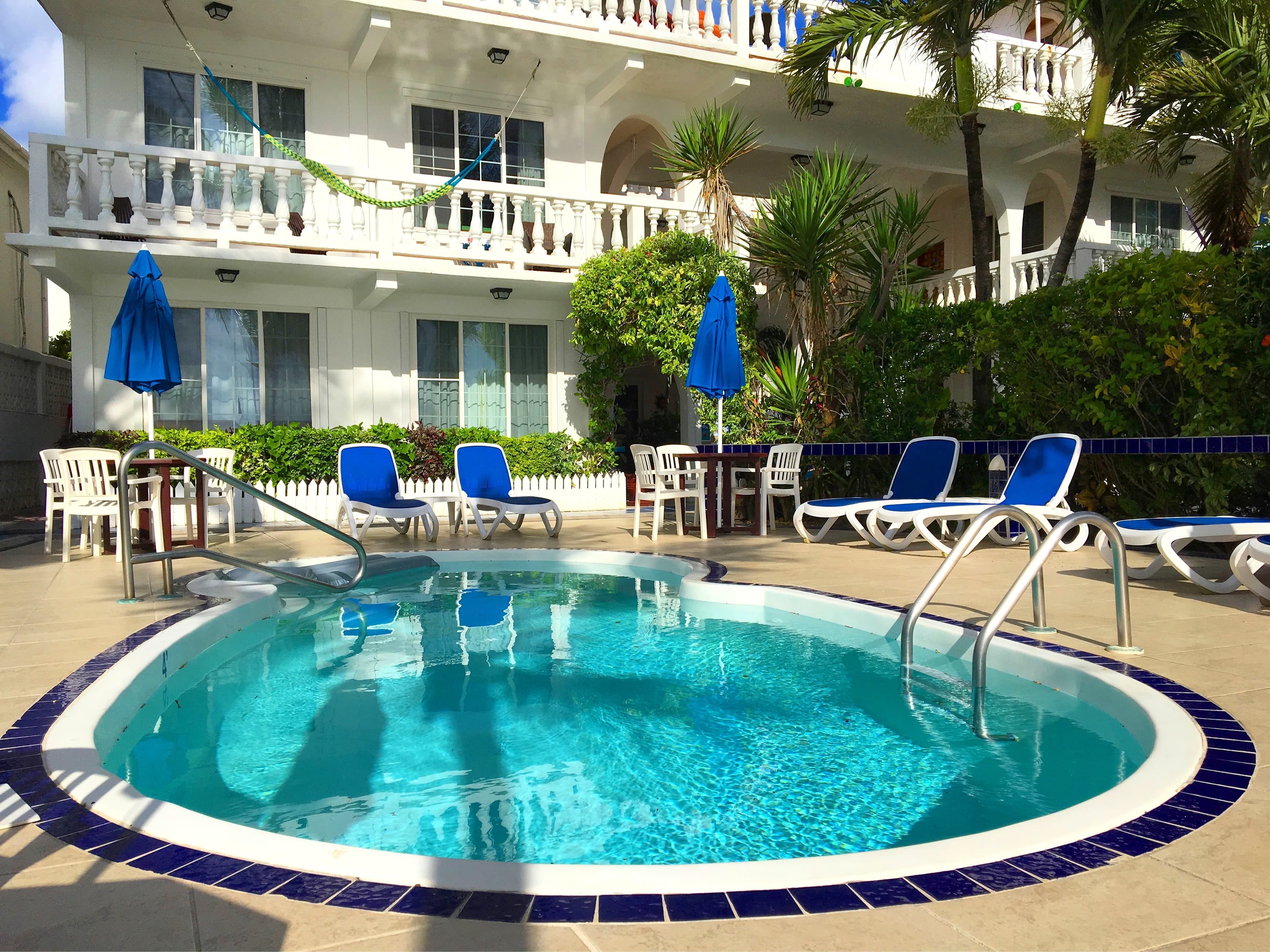 An oval swimming pool with blue and white tiling, surrounded by lounge chairs and umbrellas in front of a white building with balconies.