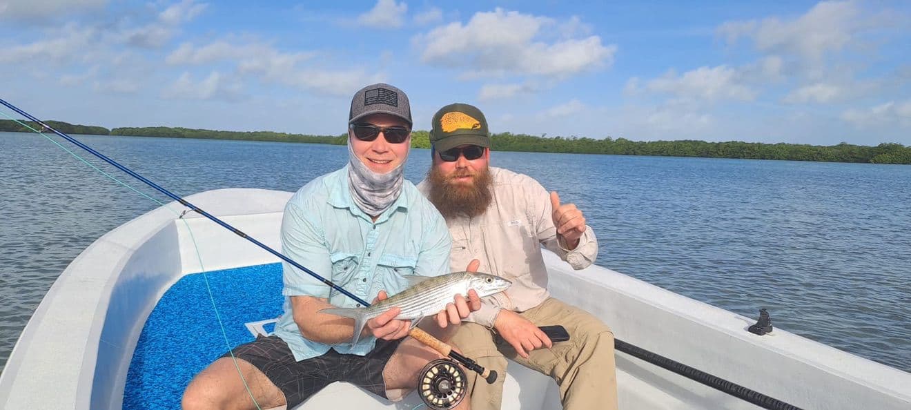 Two men on a boat proudly displaying a caught fish with a scenic water backdrop.