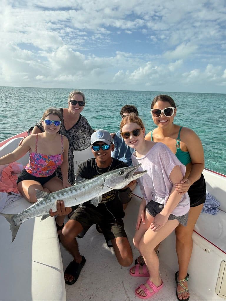 A group of five people on a boat happily pose with a large fish against a backdrop of clear blue water and sky.