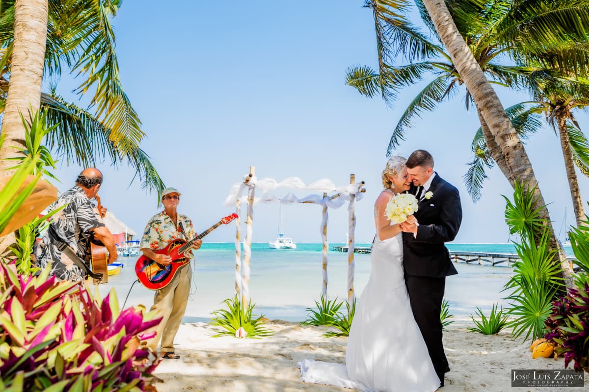A newlywed couple embraces on a sandy beach while musicians play in the background.
