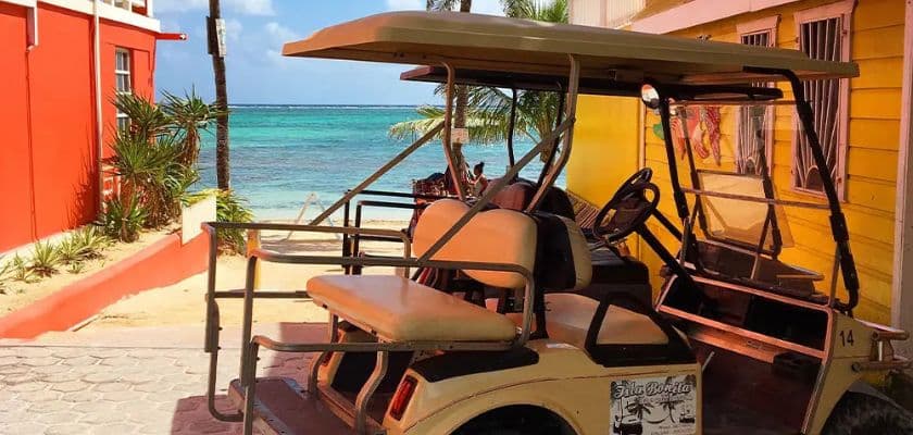 A golf cart is parked by a sandy path leading to a turquoise ocean, framed by colorful buildings.