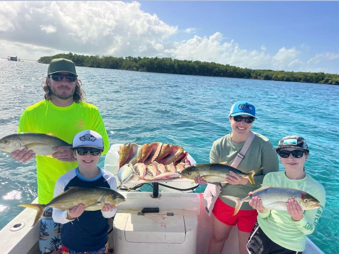 A group of four people on a boat proudly displays their catch of fish against a backdrop of clear blue water and green mangroves.