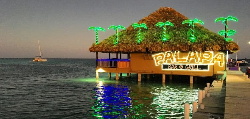 illuminated thatched-roof bar over water with palm trees and a boat in the background.