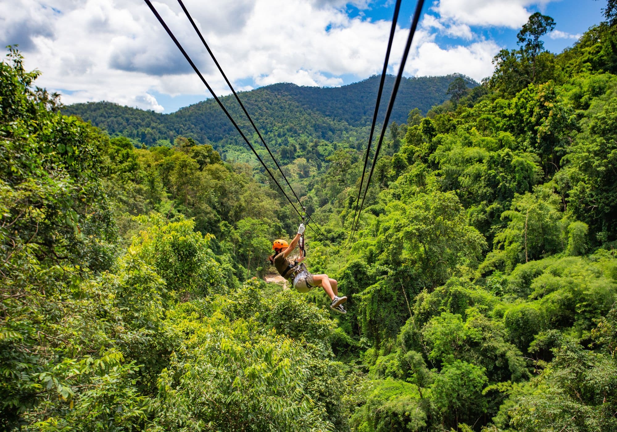 A person zip-lining above lush green forests and mountains under a bright sky.