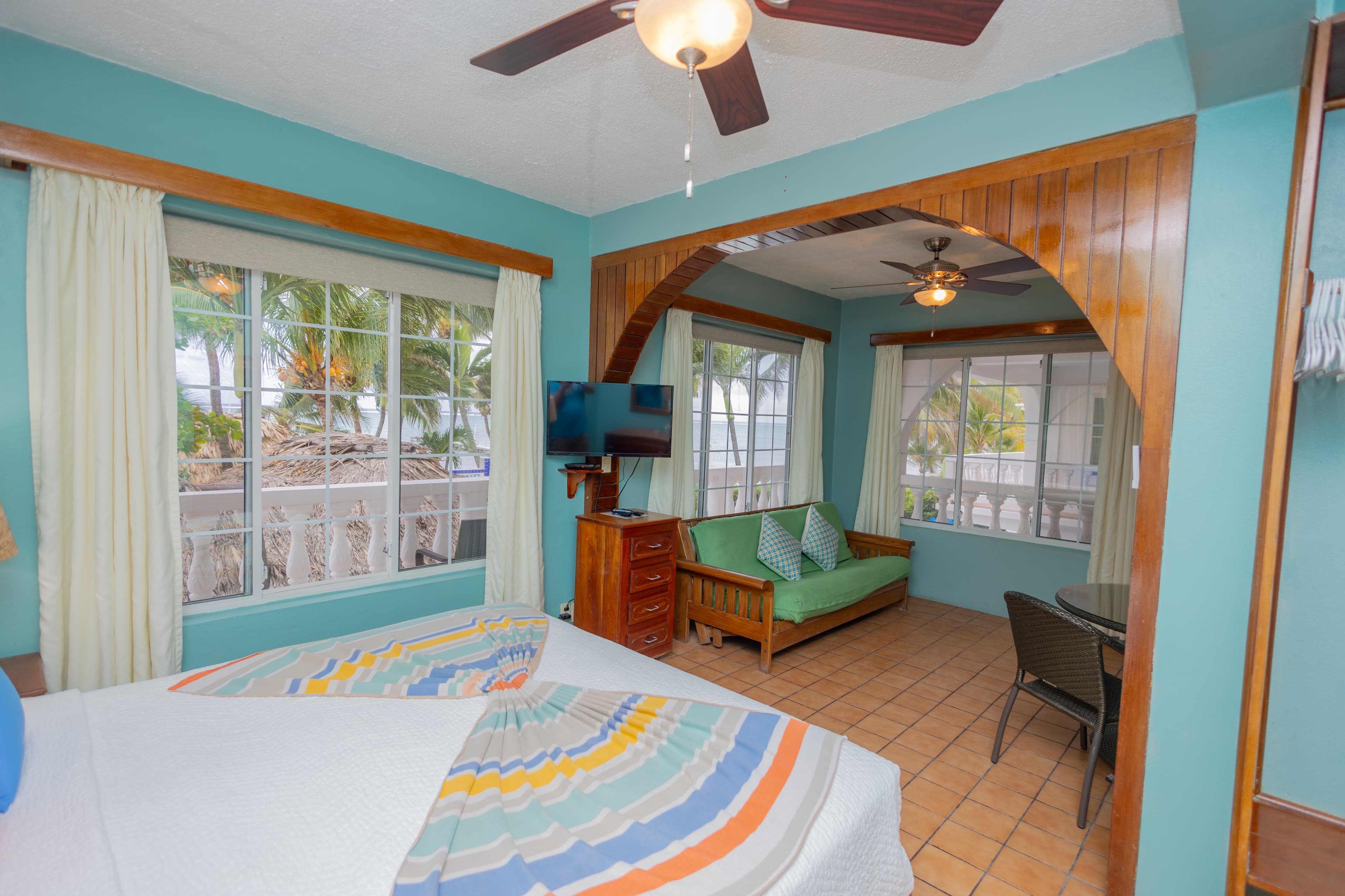 A bright, cozy guest room featuring a bed, seating area, and windows with tropical views.