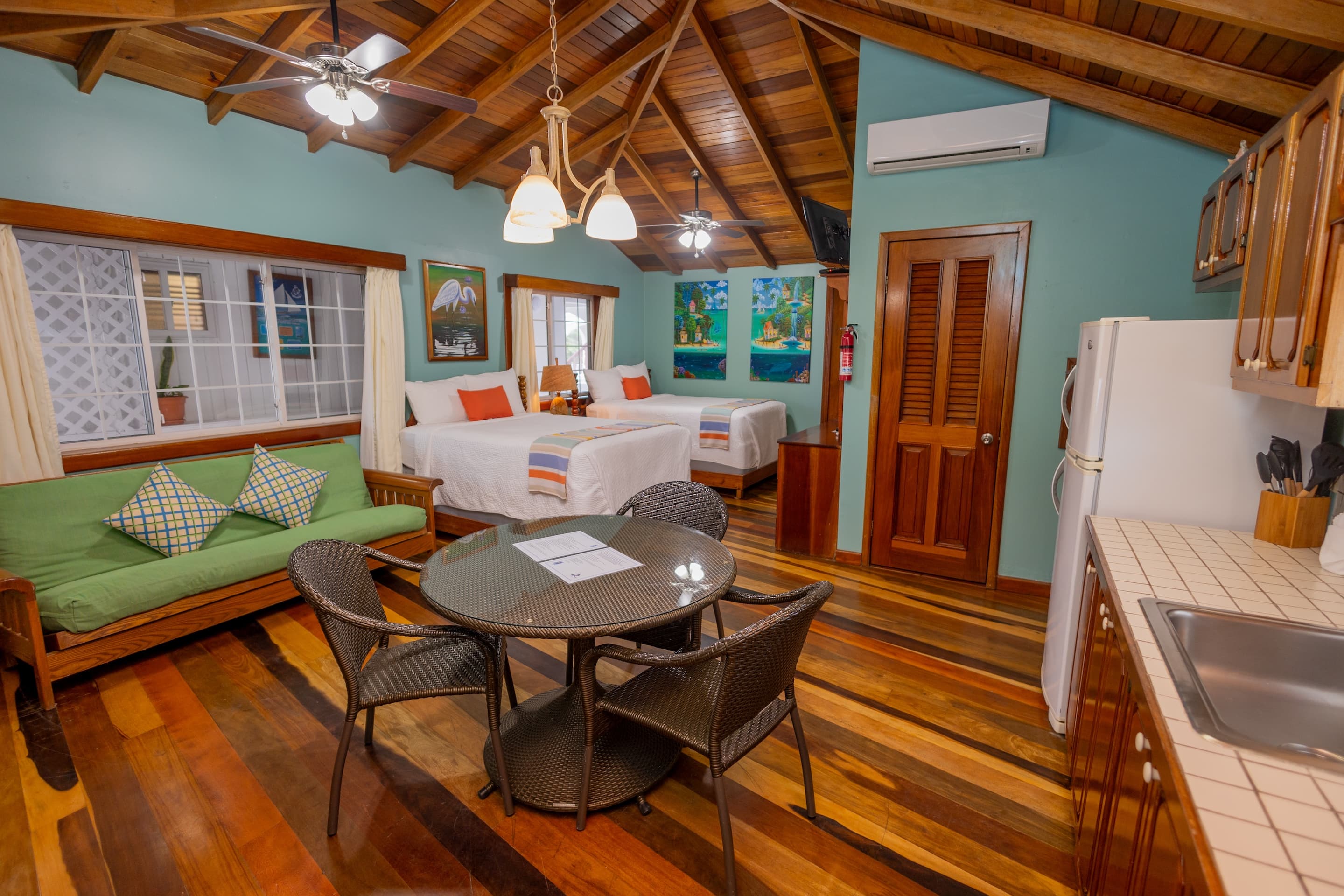 A room with a high wooden ceiling, a green futon, a round table with chairs, two beds, and a kitchenette area.