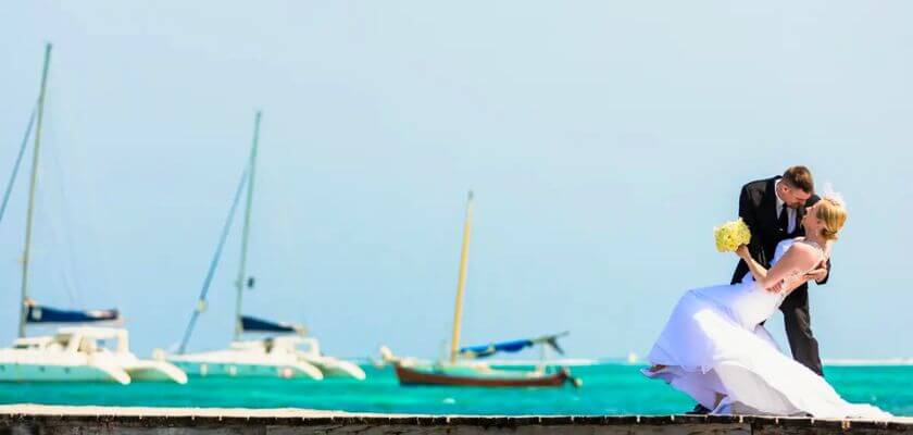 A bride and groom share a romantic moment near the ocean with sailboats in the background.