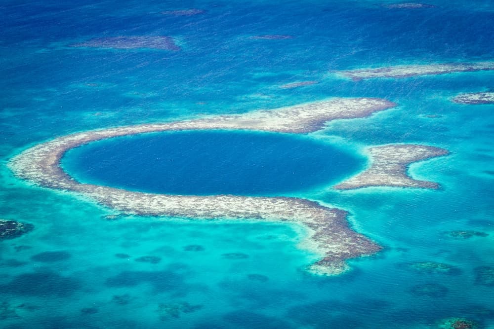 Aerial view of a circular blue hole surrounded by shallow coral reefs in turquoise water.