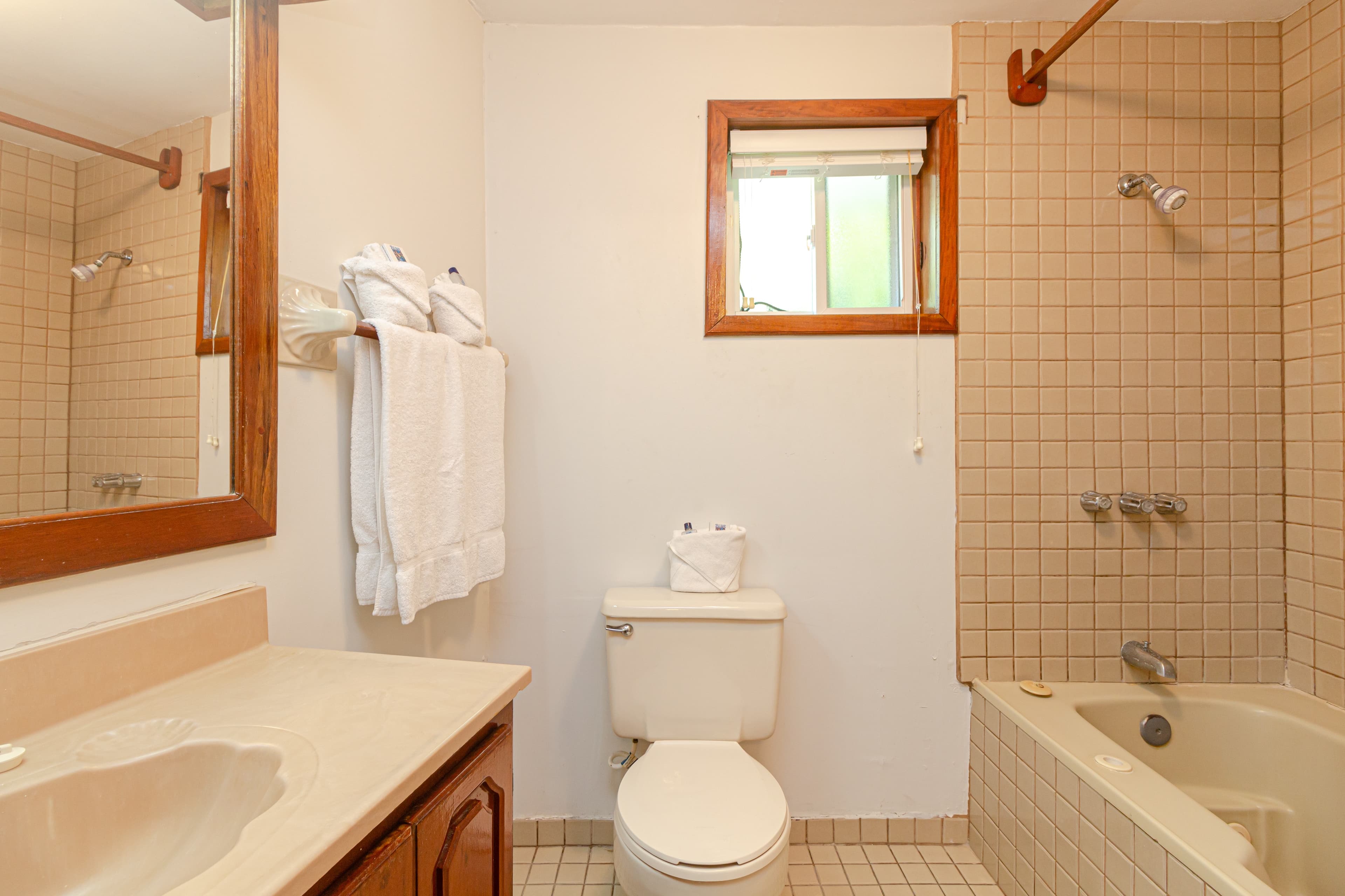 A bathroom with a bathtub and shower, a white toilet, a wooden-framed mirror above a sink, and beige tiled walls.